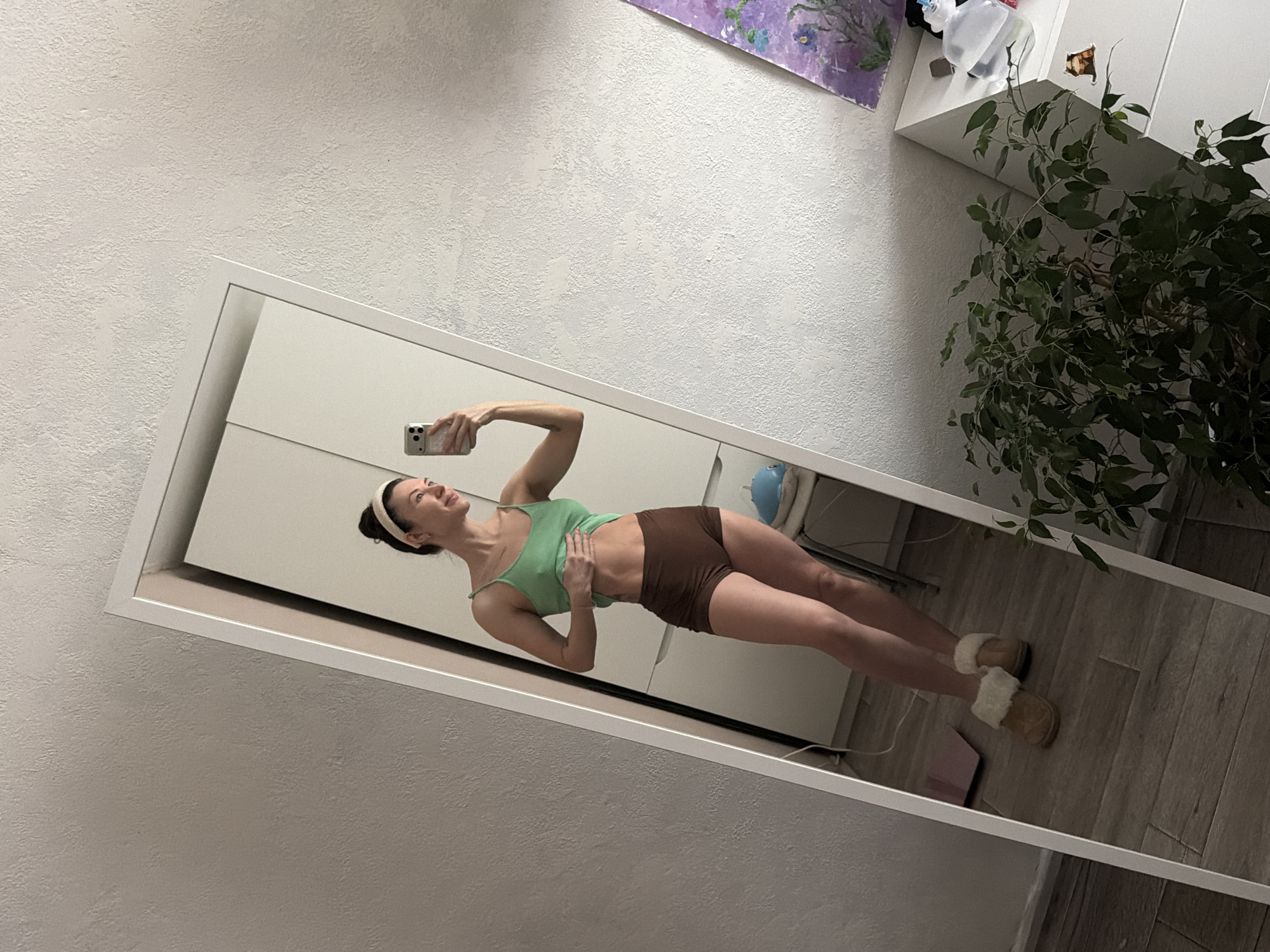 The image shows a woman reflected in a full-length mirror indoors. The woman is wearing a light green tank top, brown shorts, and fuzzy slippers. She is holding a smartphone in her right hand, seemingly taking a picture of herself in the mirror. Her left hand is placed on her stomach. She also has a white headband. The environment includes a white wall, white cabinets, a plant, a painting, and wooden flooring. The location is likely an apartment or house. The city of Perm, Russia cannot be determined from the image.