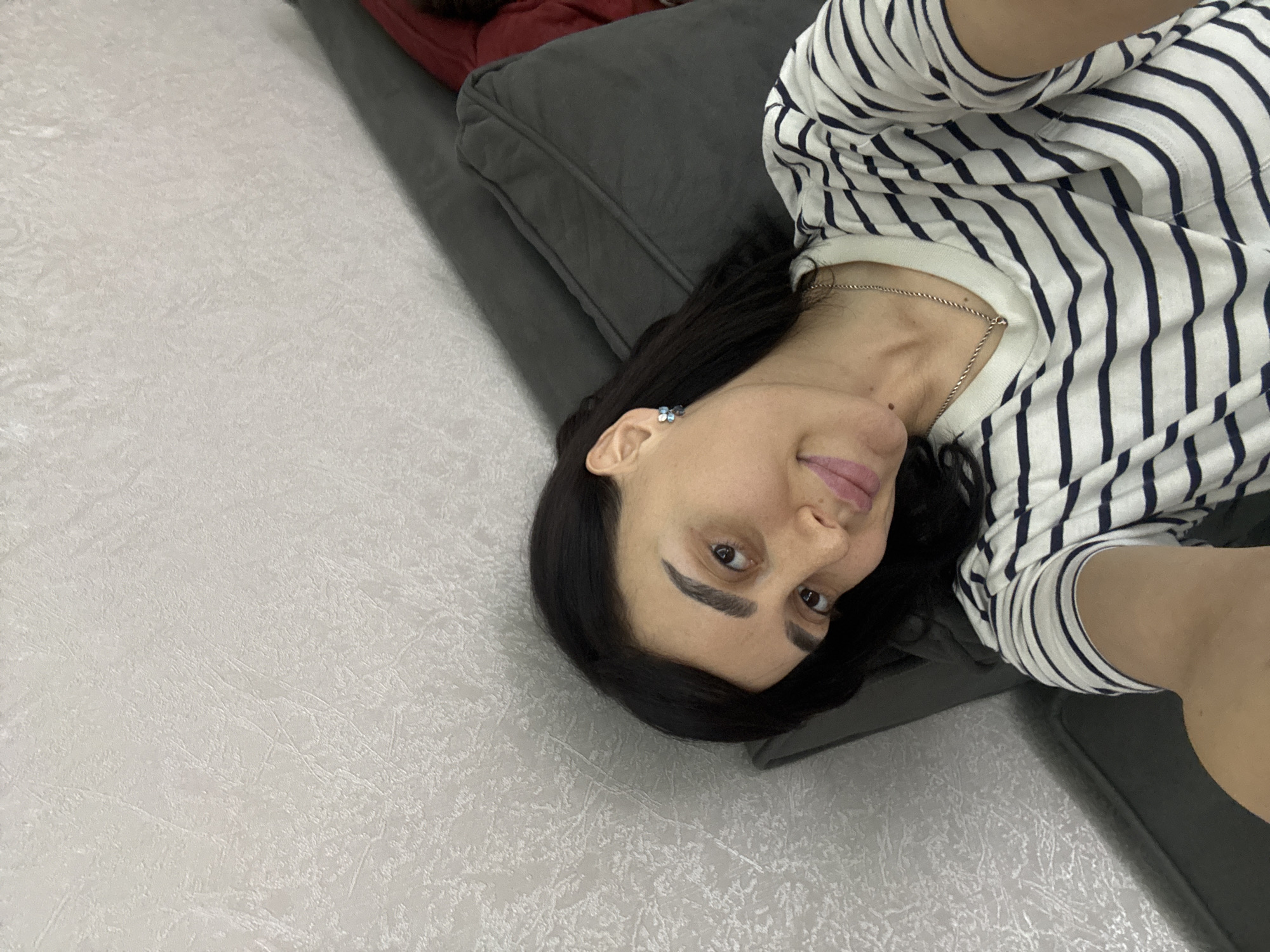 A woman with dark hair is lying on a gray sofa in what appears to be an interior space. She is wearing a striped shirt and a necklace and has an earring on her left ear. The background includes a light-colored wall. The location cannot be determined.