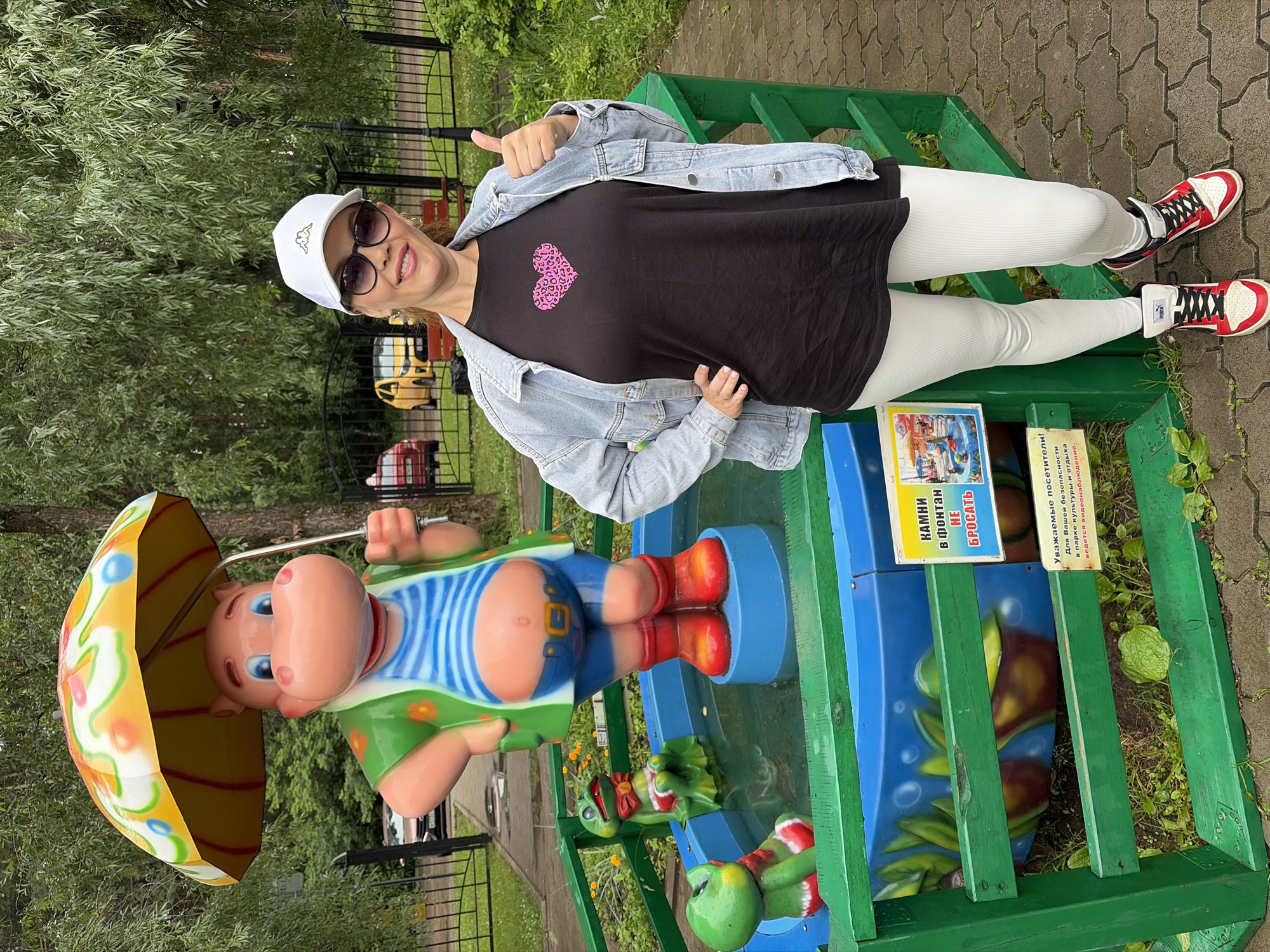 A woman with brown hair, wearing a white baseball cap, sunglasses, a denim jacket over a black t-shirt with a pink heart design, white leggings, and red and white sneakers, poses with a thumbs-up gesture next to a carousel ride. The ride features a large, cartoonish hippopotamus holding a yellow and green umbrella. The background shows lush green trees and a metal fence. The woman appears happy and is smiling. The scene is outdoors, likely in a park, given the carousel and trees. A sign on the ride reads "КАМНИ В ФОНТАН НЕ БРОСАТЬ" which translates to "DO NOT THROW STONES INTO THE FOUNTAIN". Another sign indicates "Уважаемые посетители! Для Вашей безопасности в парке культуры и отдыха ведется видеонаблюдение," which means "Dear visitors! For your safety, video surveillance is conducted in the park of culture and recreation." The weather appears to be overcast, but bright. The image seems to be taken during the daytime. The presence of a carousel and amusement park signage suggests this is a recreational area in Chaikovsky, Russia.