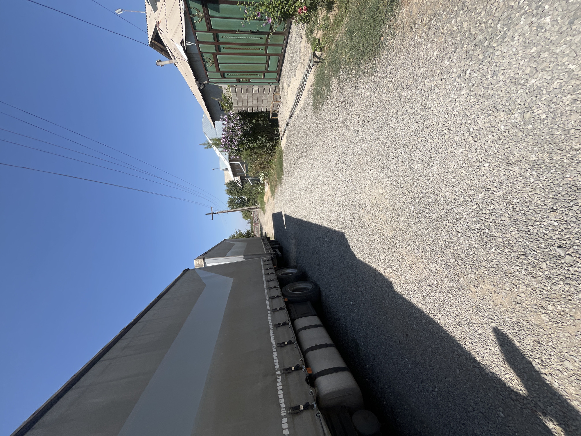 A wide-angle, low-angle shot shows a gravel road with a large, light-colored semi-trailer truck parked on the left side, casting a long shadow. The truck's tarped side is visible, and its wheels and fuel tanks are prominent. On the right side of the road, residential buildings with a distinctive green metal gate are visible. There are flowering bushes, a concrete block wall, and a wooden bench. Utility wires are strung overhead, supported by a utility pole. The sky is clear and blue. The scene appears to be in a rural or semi-urban residential area.