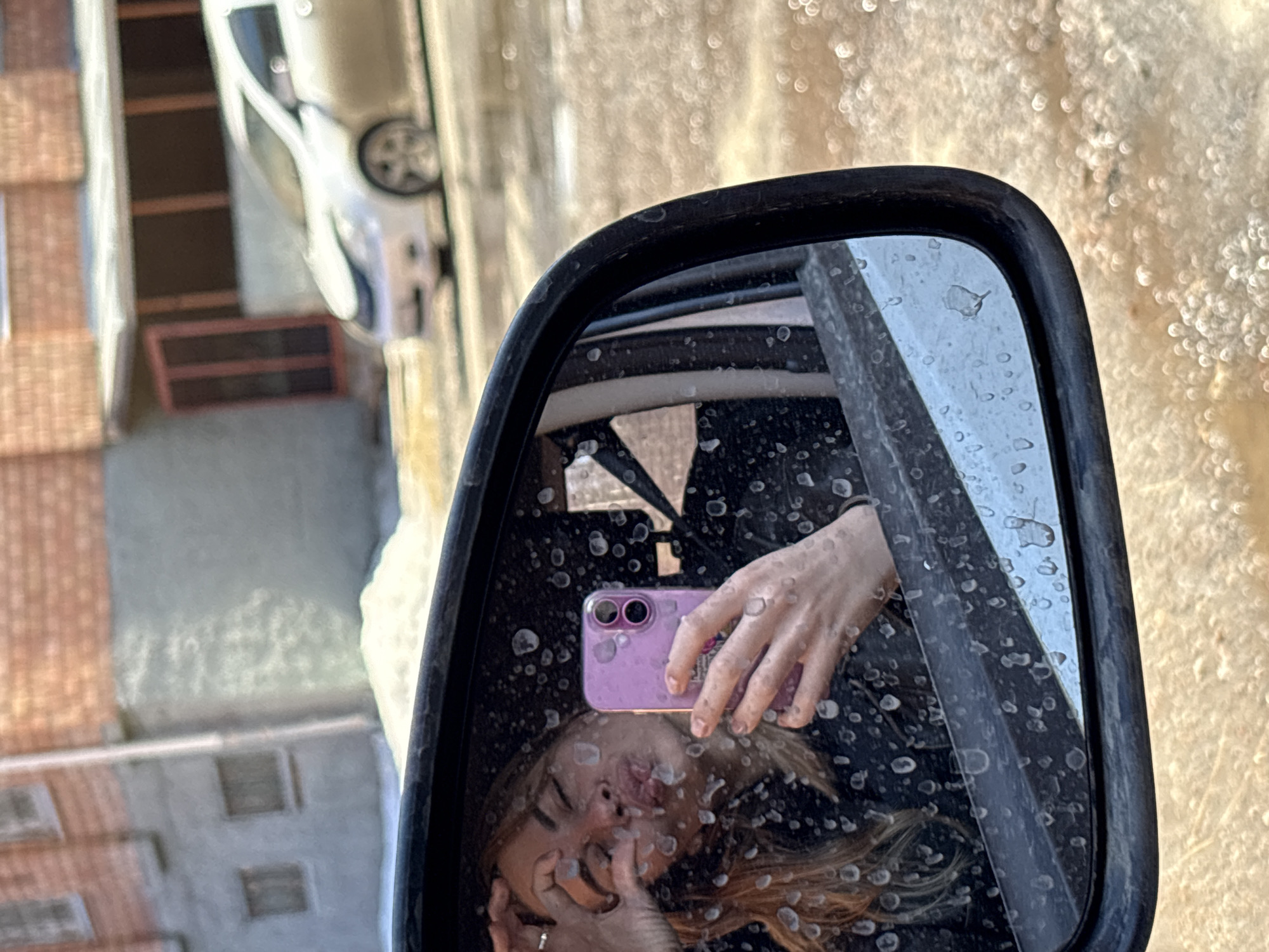 The image shows a selfie taken in a car side mirror, likely in Ust-Ilimsk, Russia. The reflection shows a young woman holding a purple smartphone, smiling at the camera. The mirror is covered in water droplets. The background reflected in the mirror includes parts of a building and a vehicle, both blurred due to the mirror's focus. Outside the mirror, the surrounding environment appears bright and indistinct, possibly due to sunlight or shallow depth of field. The orientation of the buildings suggests a street scene, typical for a city like Ust-Ilimsk.
