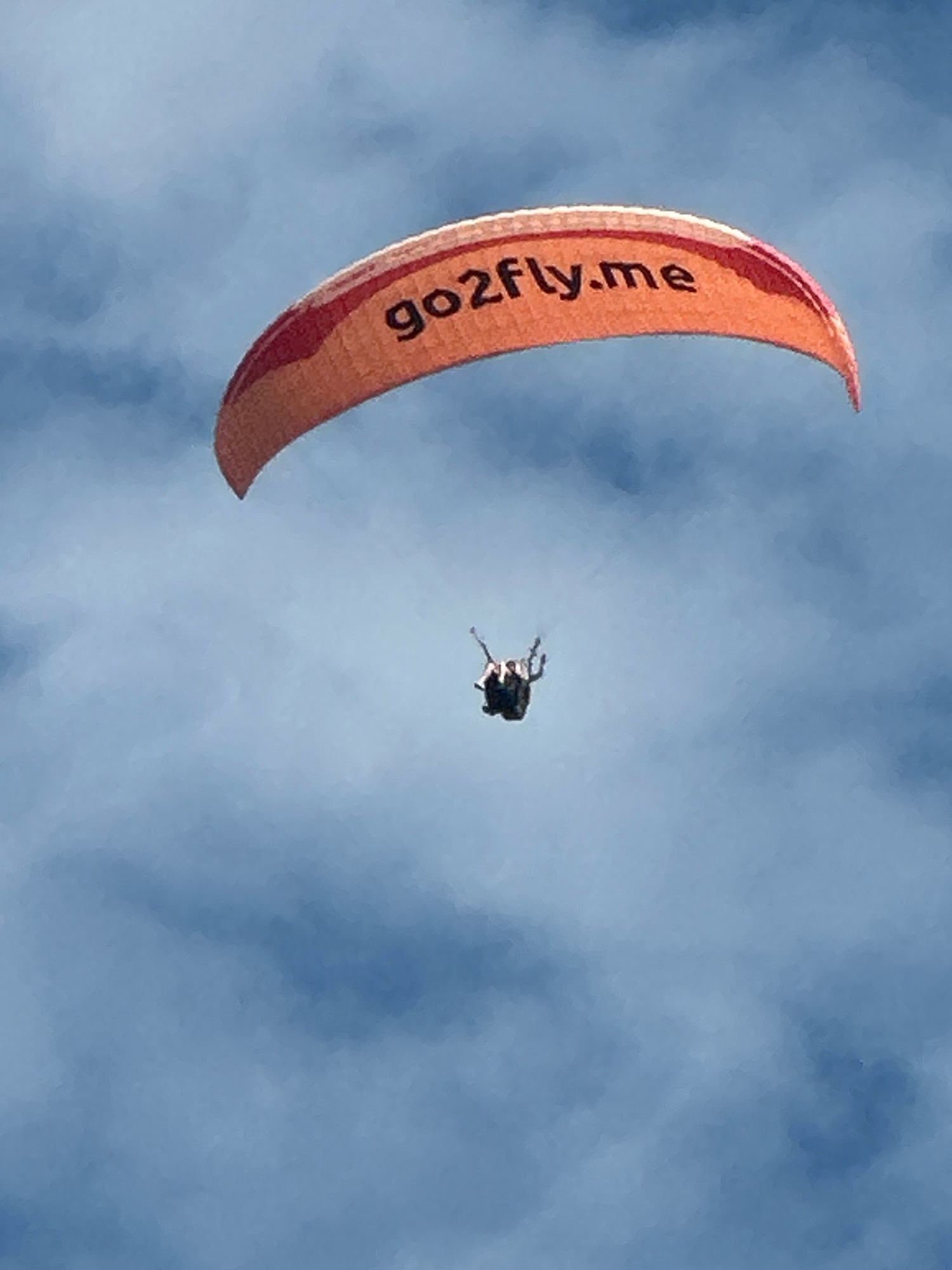 An orange paraglider with the text "go2fly.me" printed on it is soaring in a blue sky with wispy clouds. Below the paraglider, two figures are suspended in a harness, appearing to be tandem paragliding. The scene is set against a backdrop of a hazy sky, obscuring any discernible ground-level details or specific landmarks that would pinpoint a location in Boreti, Montenegro.