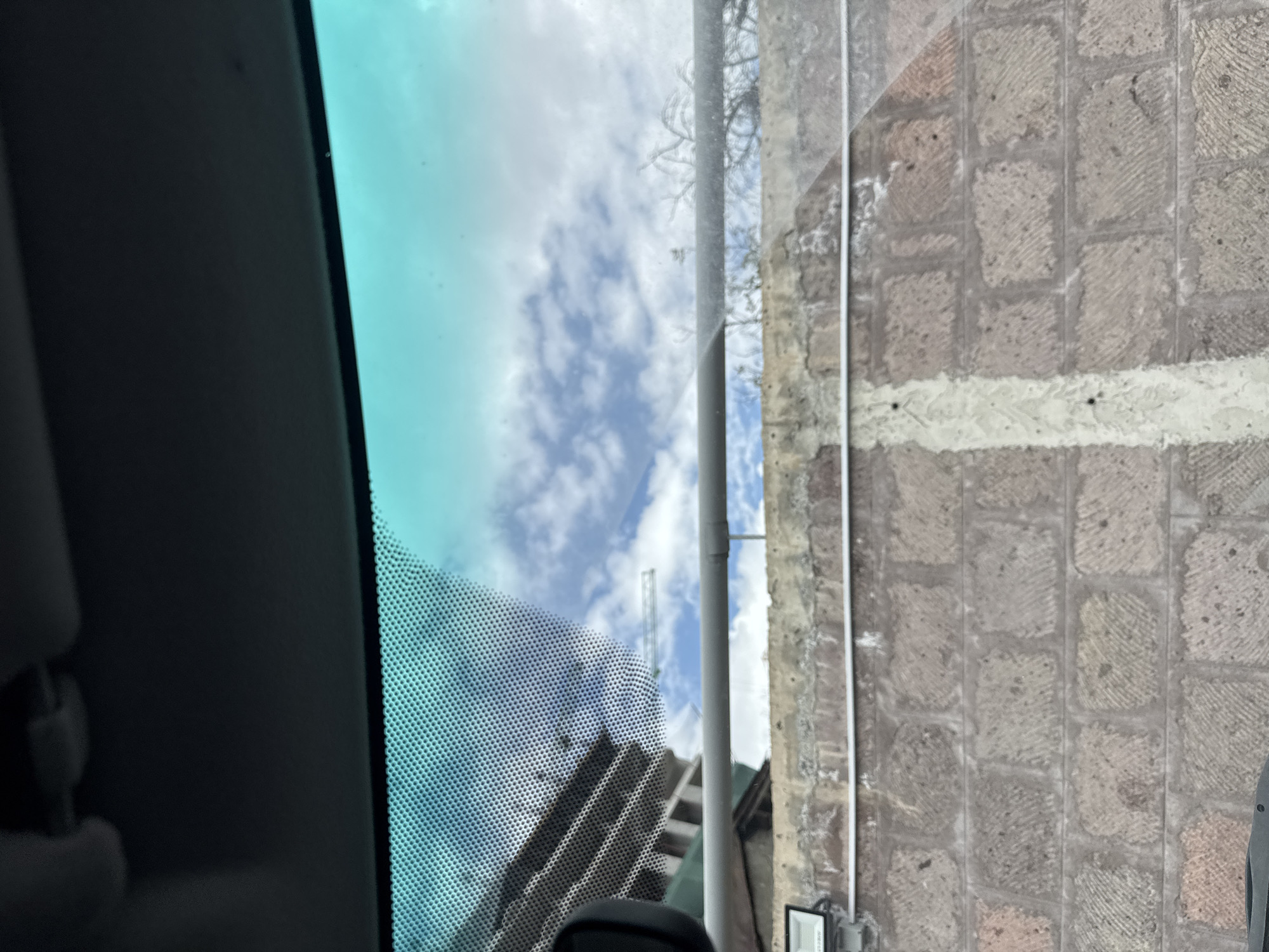 The image, taken from inside a vehicle, shows a view through a dotted windshield. The sky is visible, with patchy white clouds against a blue background. Reflections and obstructions on the windshield partially obscure the scene.
Outside the vehicle, a building with multiple floors is visible, partially obscured by a crane structure in the background. The view suggests a construction or urban environment. To the right, a wall made of stone blocks runs vertically, with a white painted line along it.
The specific location in Yerevan, Armenia is not determinable from the image alone. Further information or visual context would be required to pinpoint a precise location.