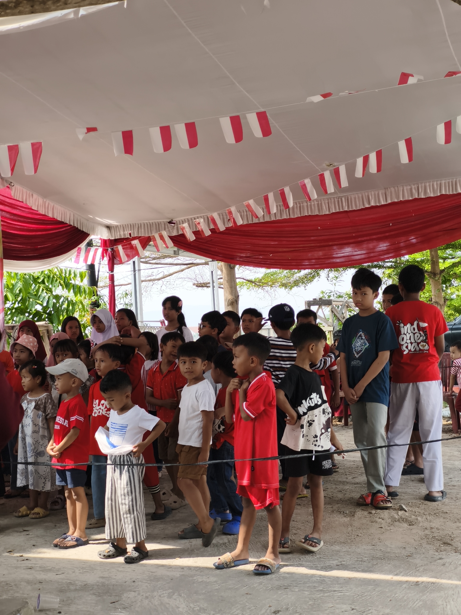 The image captures a group of children standing in a line, facing forward, likely at an outdoor event. The scene appears to be under a tent or canopy adorned with red and white bunting, characteristic of Indonesian Independence Day celebrations, as suggested by the "INDONESIA" text on one of the children's shirts. The children are dressed in casual attire, with many wearing red and white, further reinforcing the patriotic theme. Some children are holding white pieces of paper, and one boy in the foreground is adjusting his striped pants. The ground is a rough, sandy surface, and in the background, lush green foliage and trees are visible, hinting at a tropical environment, consistent with Bandar Lampung, Indonesia. The lighting suggests it is daytime.