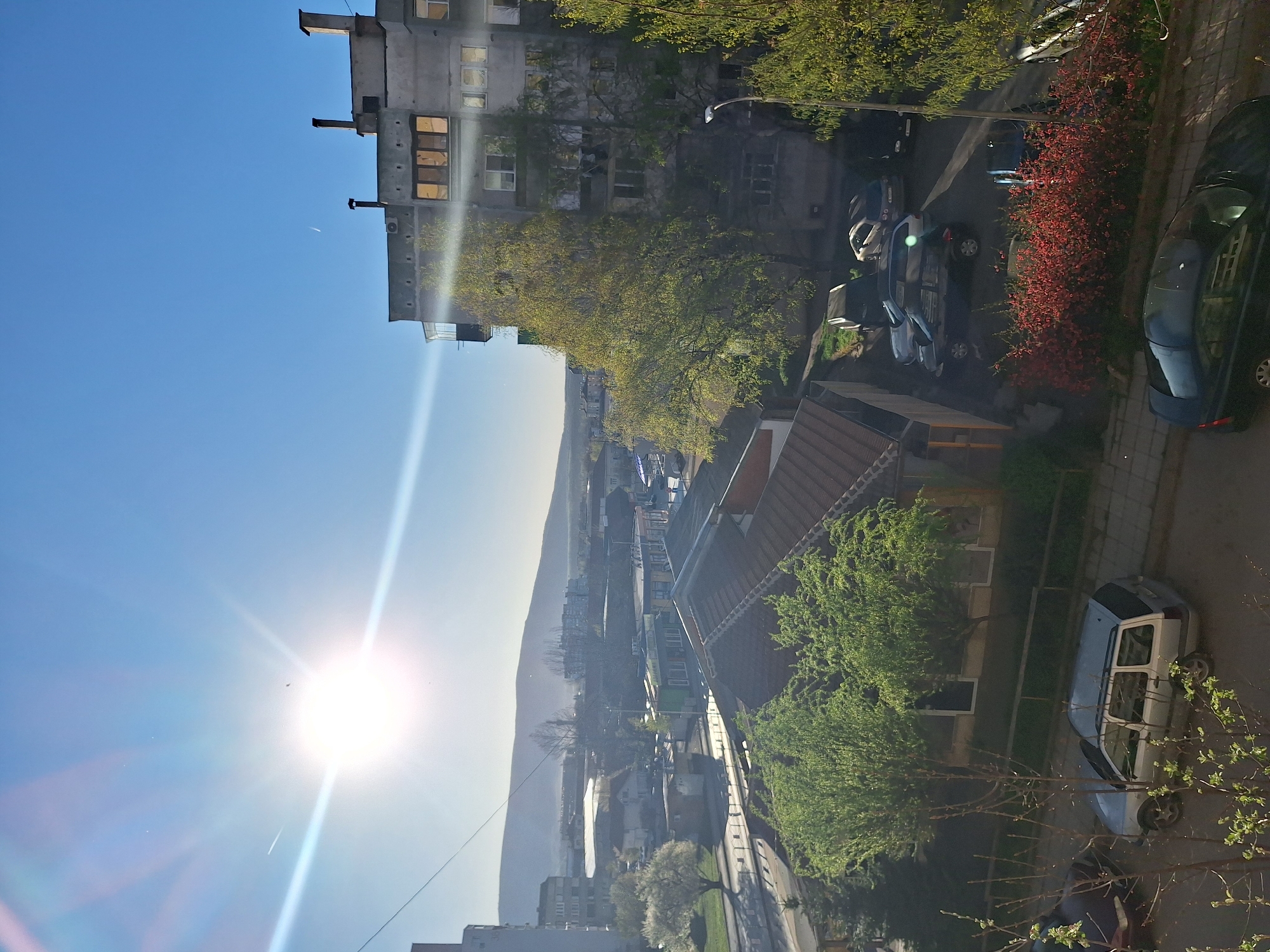 The image depicts an elevated view of an urban area during daylight. The sun shines brightly from the left side of the frame, causing lens flare. A multi-story concrete building is on the right. A tree with yellow leaves is near the building. Several parked cars are along the street. A bush with red leaves is also visible. A mountain is in the distance. Other buildings, streets, and greenery are in the middle ground. A white vehicle is partially obscured by trees in the foreground. The location is Unknown, Unknown.