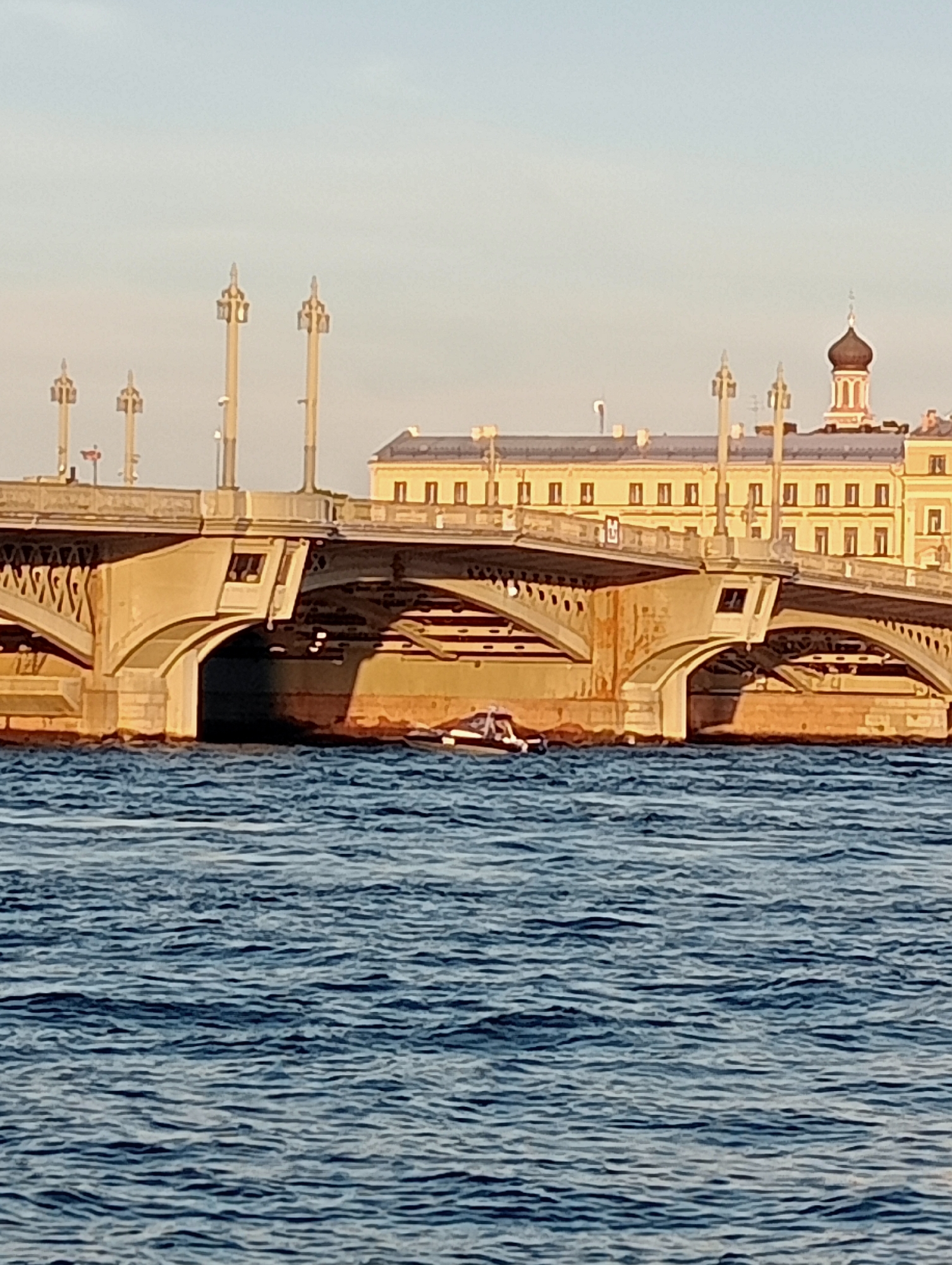 The image shows a wide, stone arch bridge spanning a body of water in Saint Petersburg, Russia. The bridge has elaborate lamp posts along its top. In the background, a light-yellow building with a dark-domed tower is visible behind the bridge. The water in the foreground is rippled and blue. A small, dark boat with a white stripe is partially visible under the bridge arch on the left side of the image. The sky is a pale blue, indicating daylight, possibly late afternoon due to the warm lighting on the bridge.
