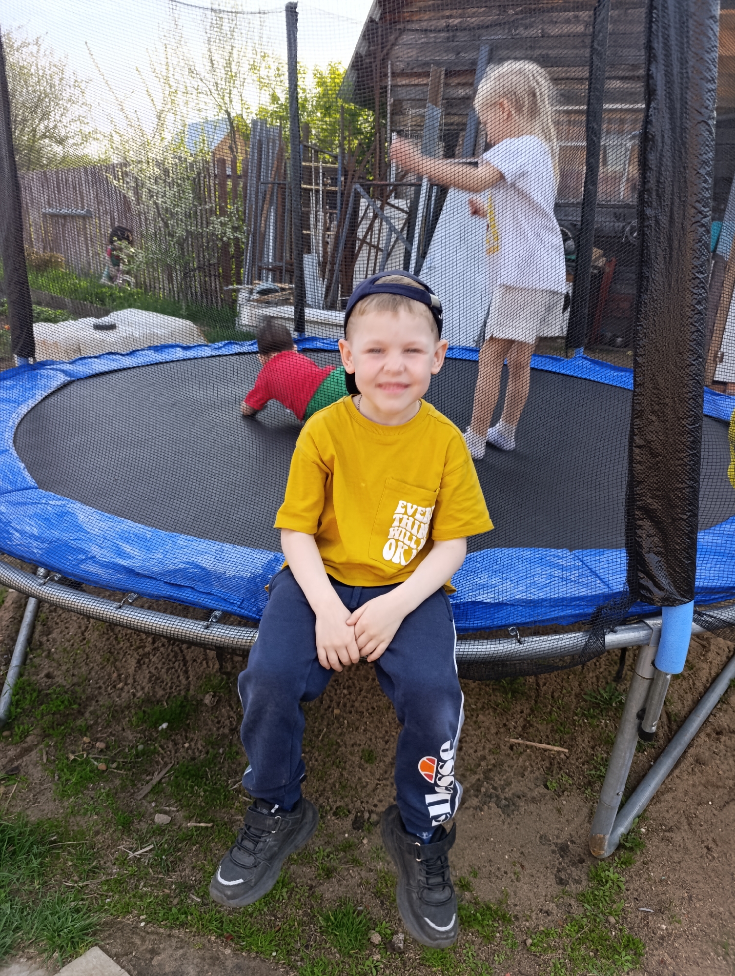 A young boy sits on the edge of a blue and black trampoline with a safety net, smiling at the camera. He wears a yellow t-shirt, dark blue pants with a white stripe and "Ellesse" logo, a dark blue baseball cap, and black sneakers. Two other children are on the trampoline. One is wearing a white t-shirt and white shorts and is standing near the safety net. The other is wearing a red shirt and green pants and is on all fours in the back. The trampoline is located in a yard with patchy grass and dirt. A wooden fence and outbuildings are visible in the background. This image was taken in Chaikovsky, Russia.