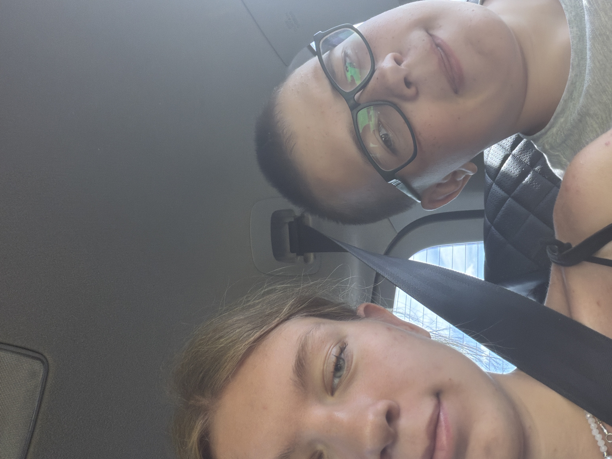 Two individuals, a female and a male, are captured in a close-up, selfie-style photograph within a vehicle. The female, positioned on the left side of the frame, has light blonde hair and is looking towards the camera with her mouth slightly ajar. She is wearing a necklace with white beads. The male, on the right, is wearing glasses with black frames and a grey t-shirt. His expression is neutral, with his head tilted slightly. A black seatbelt is visible across both individuals, indicating they are likely passengers. The background shows the interior of a car, including a portion of the headliner and a window reflecting the sky. The scene is set during daylight hours. The image is not in Novoe Feizullovo, Russia.