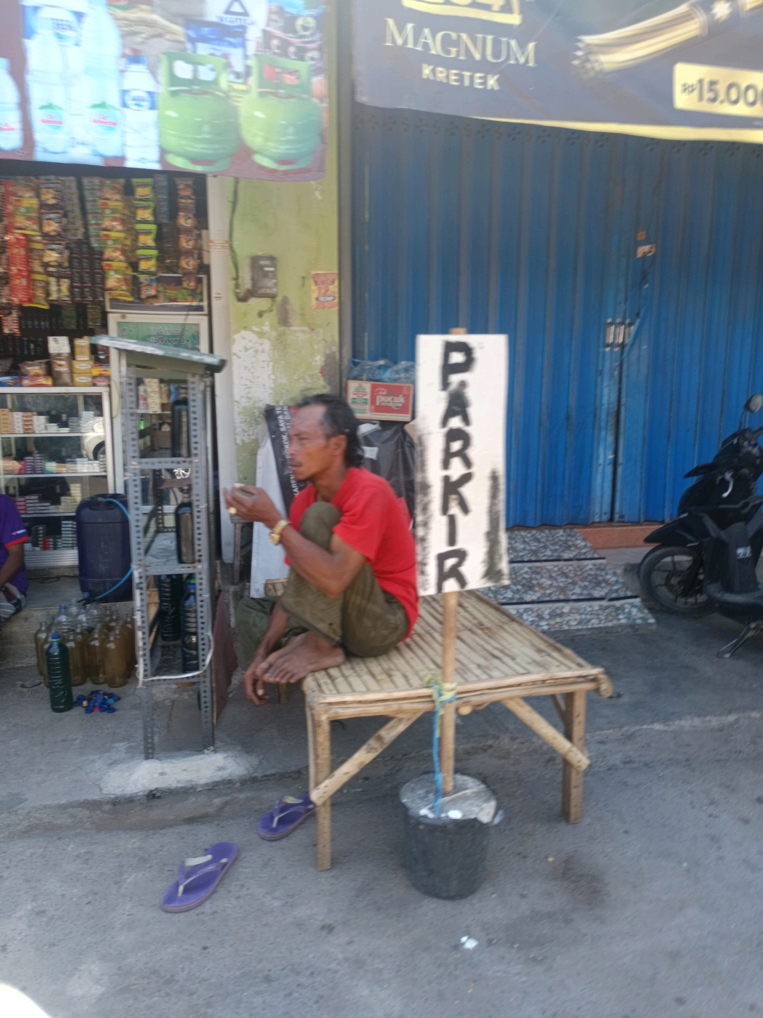 The image depicts a street scene in Blega, Indonesia. A man wearing a red shirt and green pants sits on a bamboo bench near a blue metal shuttered storefront. A sign that reads "PARKIR" is propped up on a wooden stick in front of him. To his left, a metal shelf unit displays bottles, some of which contain liquid. In front of the shelf unit are several glass bottles filled with a dark liquid and some blue bottle caps. A dark blue plastic barrel is also visible to the left of the shelf. To the right of the bench, a black motorcycle is parked. Purple flip-flops are lying on the ground in the foreground. The ground appears to be made of concrete. A large blue tarp or corrugated metal structure forms the background.