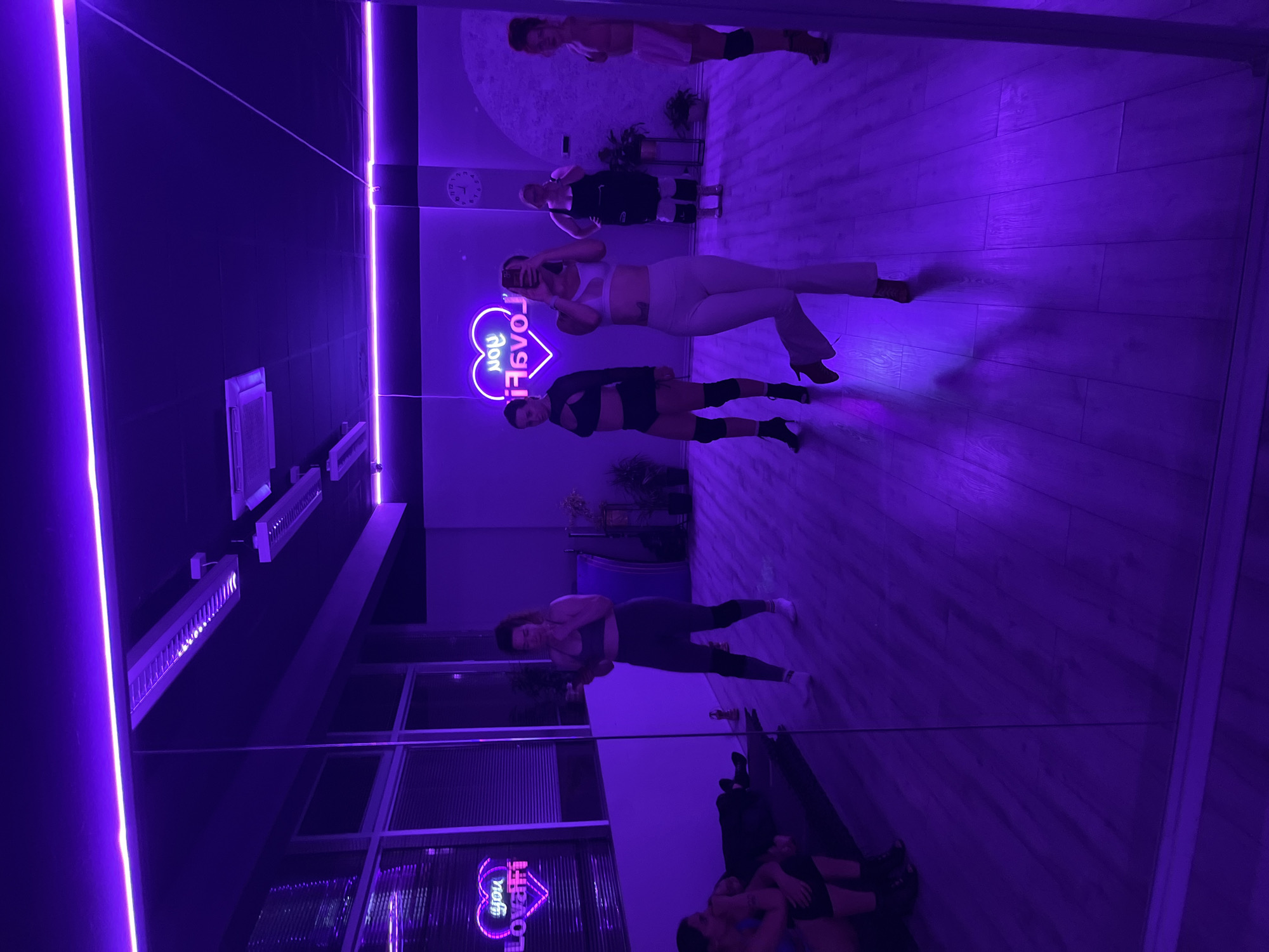 The image shows a dance studio in Budva, Montenegro, illuminated with purple neon lighting. Three women are standing, seemingly posing or taking a photo in front of a large mirror. One woman is holding a mobile phone up, likely taking a photo. The floor is wooden. Reflected in the mirror are other people further back in the studio. There is a neon sign that reads "Love You." Windows are visible along the wall.
