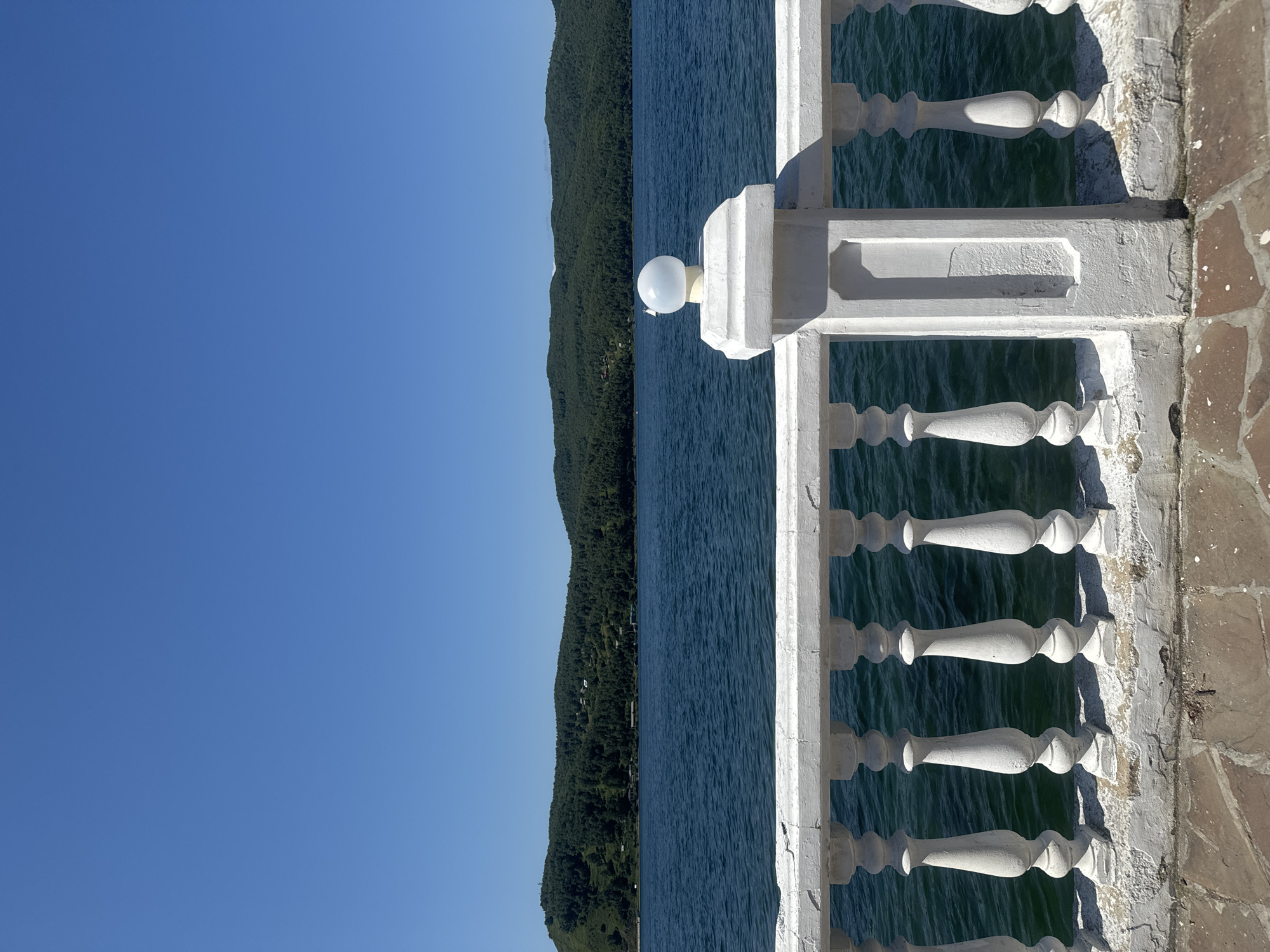 A waterfront view of a calm, blue lake with forested hills in the background is framed by a white, ornate balustrade. The balustrade, made of concrete and painted white, shows signs of age and wear with chipped paint and visible cracks along its base. The balustrade features a repeating pattern of turned balusters, topped by a horizontal railing. A decorative post with a spherical, frosted glass light fixture sits prominently in the right portion of the balustrade. The ground in front of the balustrade is paved with irregular-shaped, reddish-brown stones. The sky above is a clear, vibrant blue. The scene is likely located in Listvyanka, Russia, a village situated on the western shore of Lake Baikal.