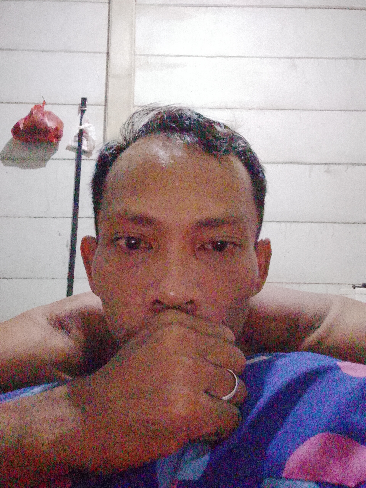 A close-up, slightly grainy selfie of a shirtless man with short, dark hair and a light complexion. He is lying down, resting his weight on his elbows and forearms. His right hand is raised to his mouth, partially covering his lower face. A silver ring is visible on his ring finger. The background consists of a white wall with horizontal paneling. A red plastic bag and a white cloth or bag hang from a rod or pole in the background. The man is lying on a blue and pink patterned bedspread. The location is Bukit Ketok, Indonesia.