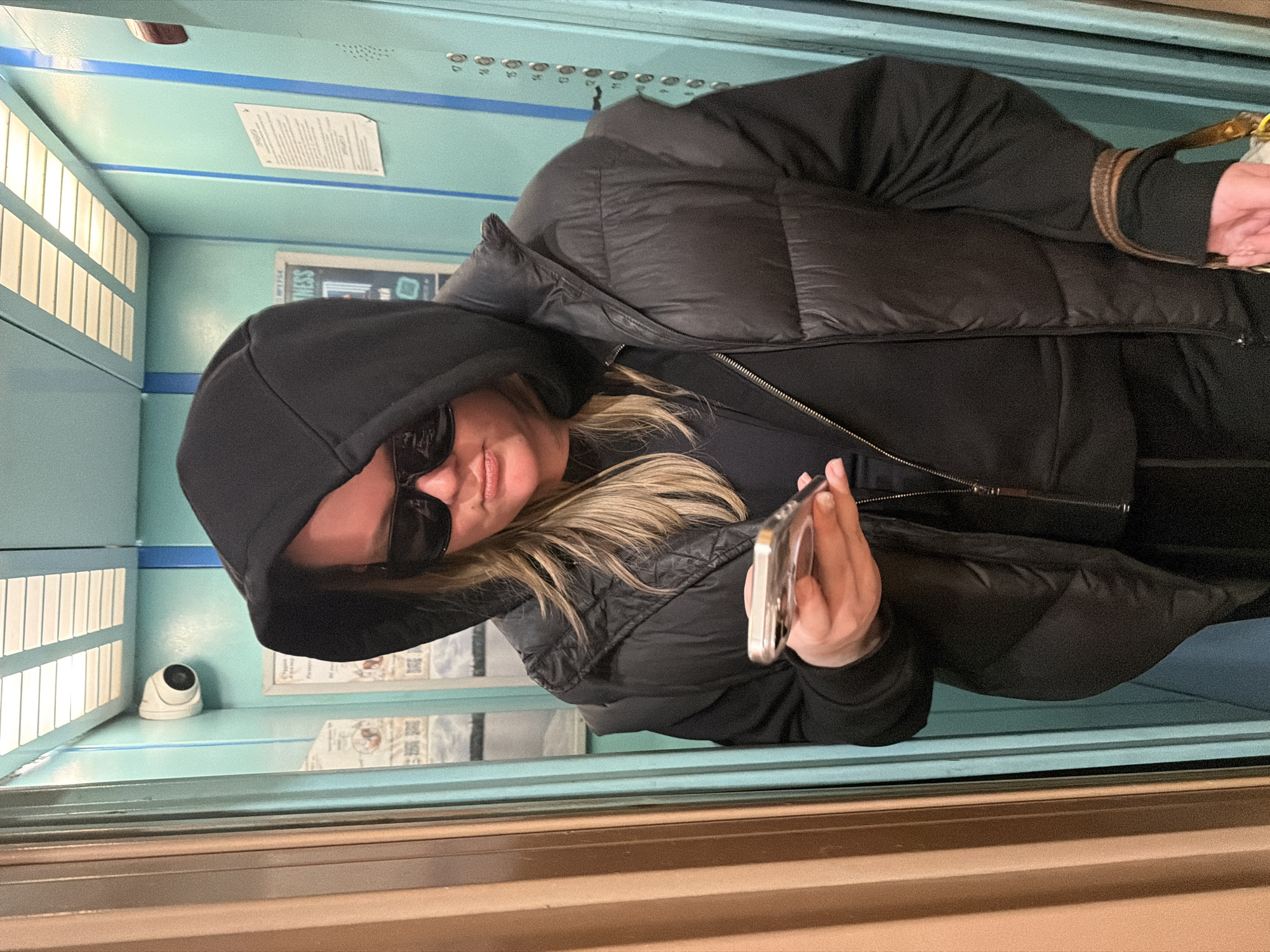 A woman is in an elevator, holding a cell phone. She is wearing a black hooded sweatshirt, sunglasses, and a black puffer jacket. The elevator has a row of lights along one wall and a security camera. There are posters on the wall behind her and elevator buttons above the posters. The scene is in Saint Petersburg, Russia.