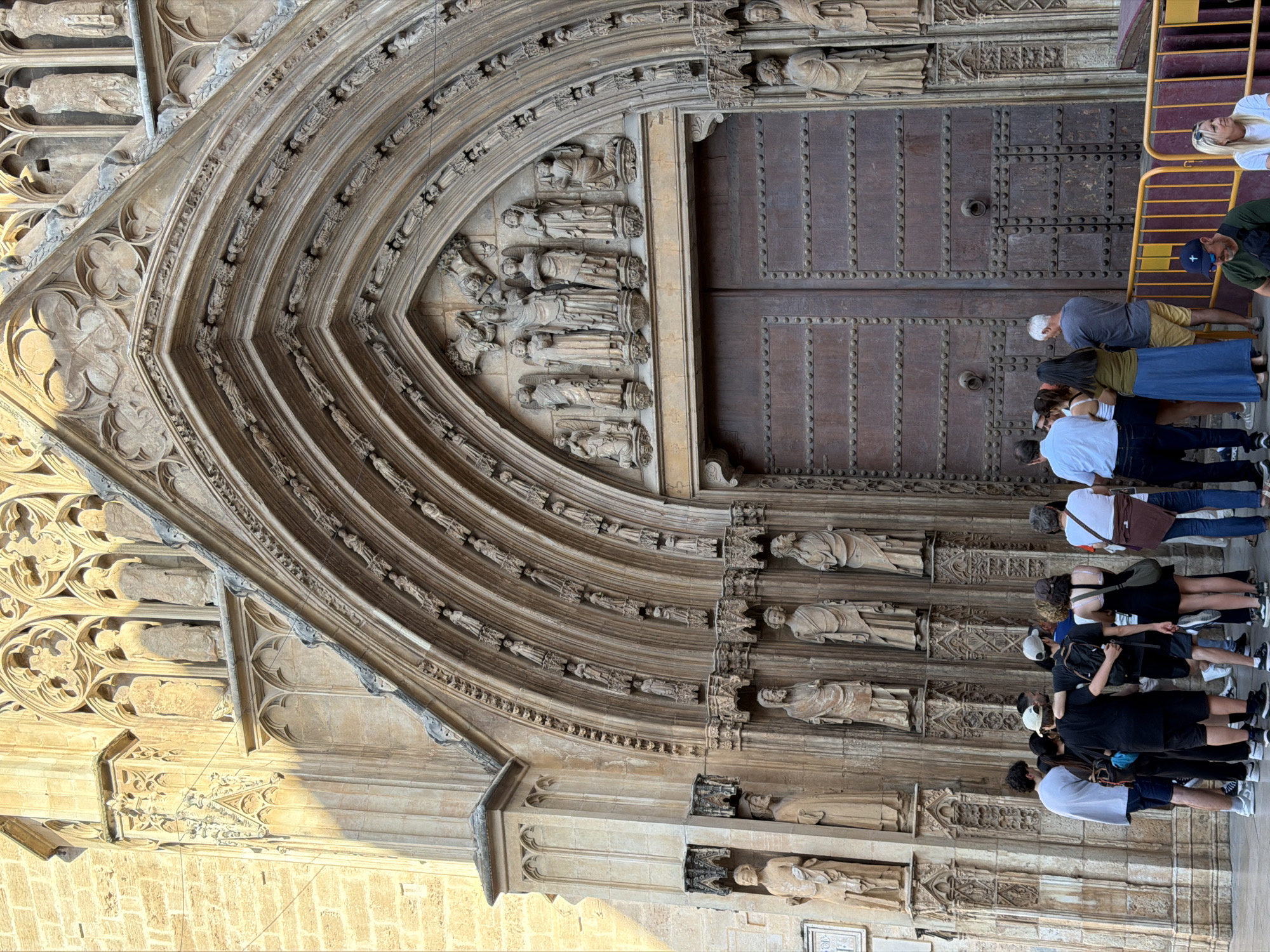 This is a medium-angle shot of a group of tourists gathered in front of a large, ornate church entrance. The church's facade is made of light-colored stone and features intricate Gothic architecture, including pointed arches, detailed carvings, and numerous statues of religious figures. The main entrance is a massive wooden door with prominent metal rivets. The sunlight casts shadows across the facade, highlighting the textures and depth of the carvings. The tourists, a mix of men and women of various ages, are casually dressed and appear to be observing the architecture. Some are looking at the door, while others are gazing upwards at the sculptures. One man has his arm around another man's shoulder, suggesting a friendly interaction. A yellow metal barrier is present in the foreground, separating the tourists from the building. The ground in front of the church is paved.