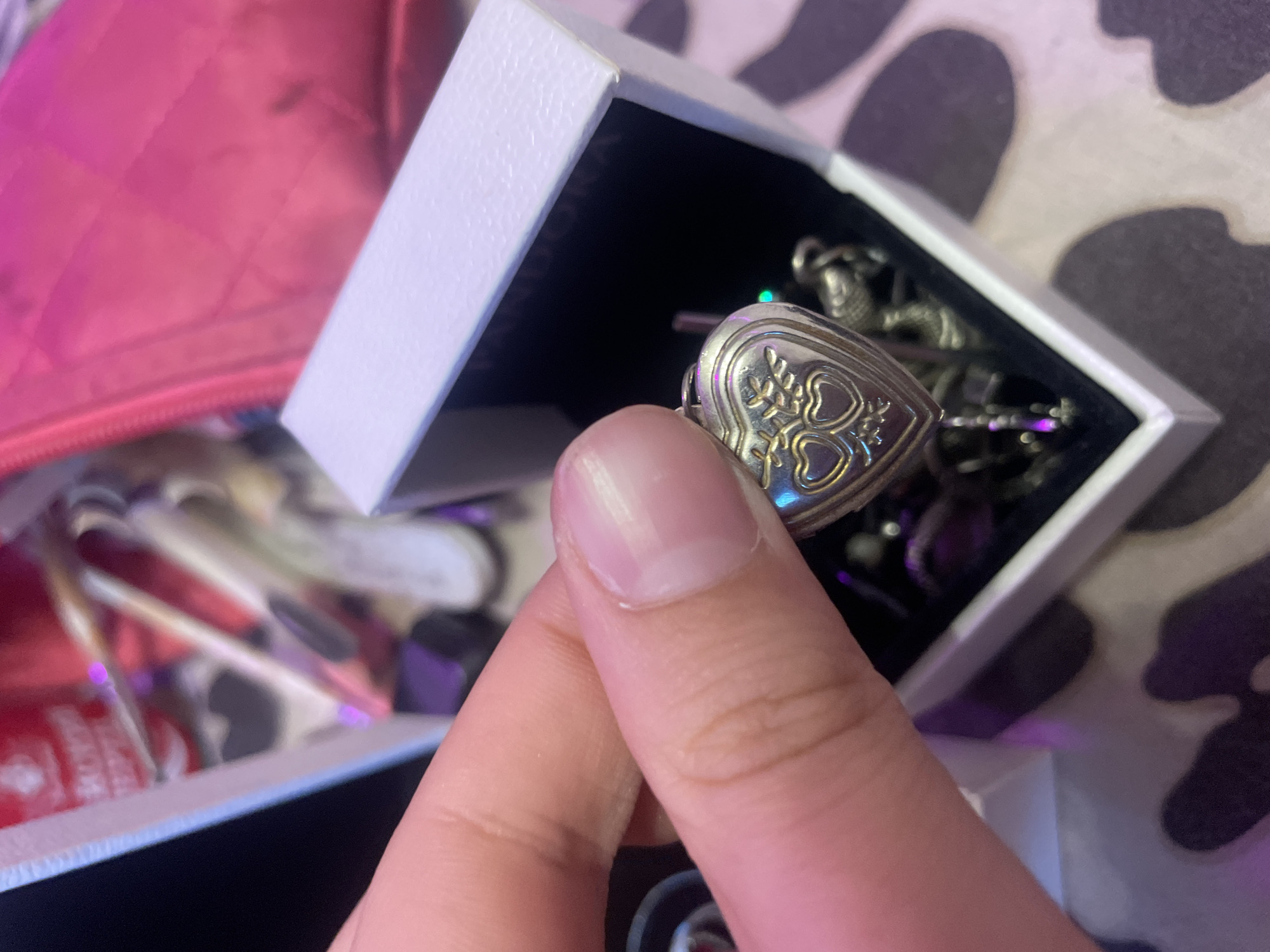 A hand holds a heart-shaped locket with two smaller hearts inside and wings on either side. The locket is silver with gold accents and is placed in an open white jewelry box. The background shows a pink quilted bag and a blurry red tube. The scene is lit with purple and green lights, suggesting an indoor setting. No specific location in Ladozhskoe Ozero, Russia can be determined from the image.