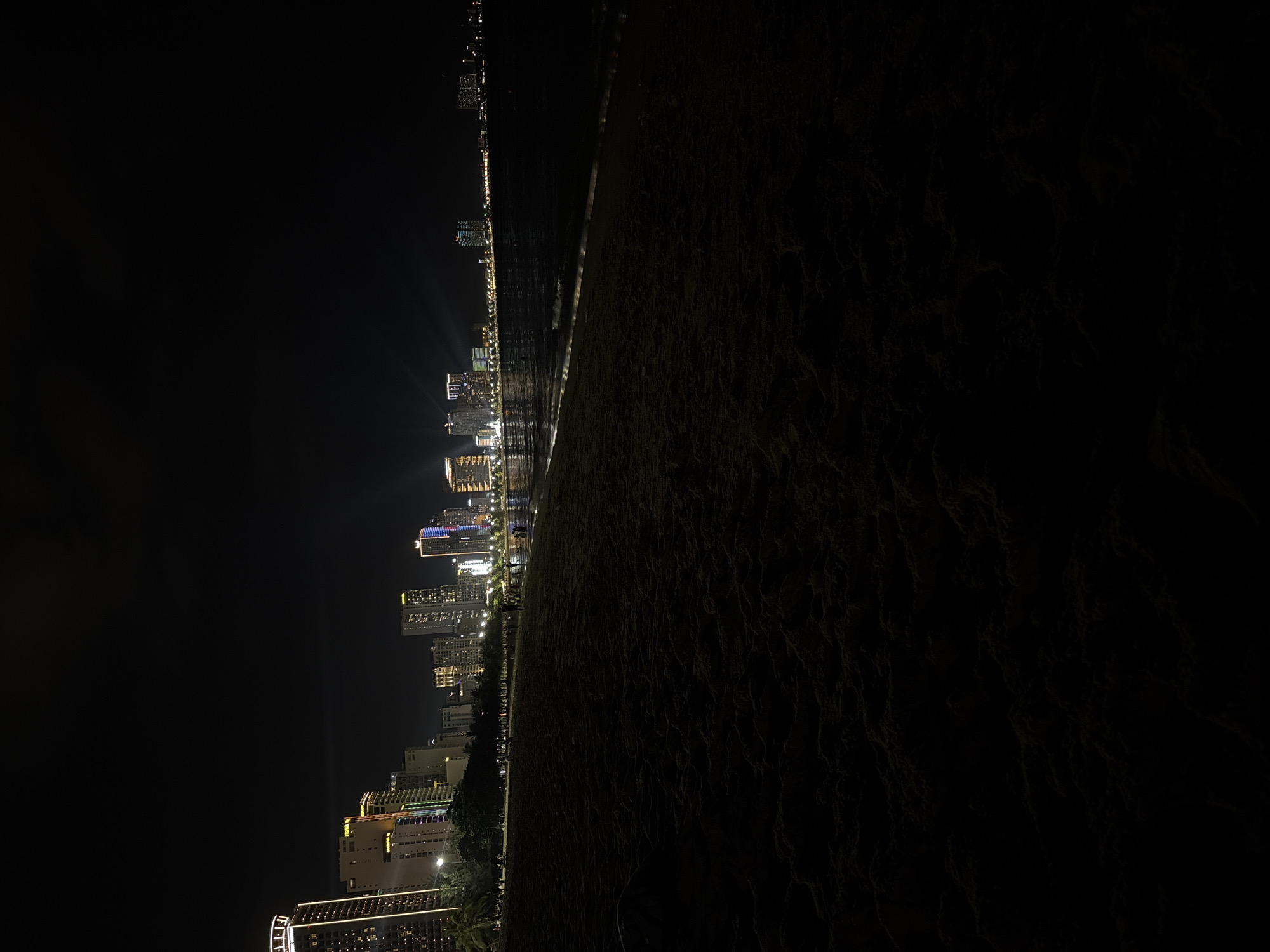 At night, a coastal cityscape stretches out along the Nha Trang Ward, Vietnam shoreline, marked by illuminated buildings and the adjacent dark beach. The city's lights reflect on the water's surface, and the coastline is visible in the distance. The photograph is taken from ground level. There are no observable individuals present.