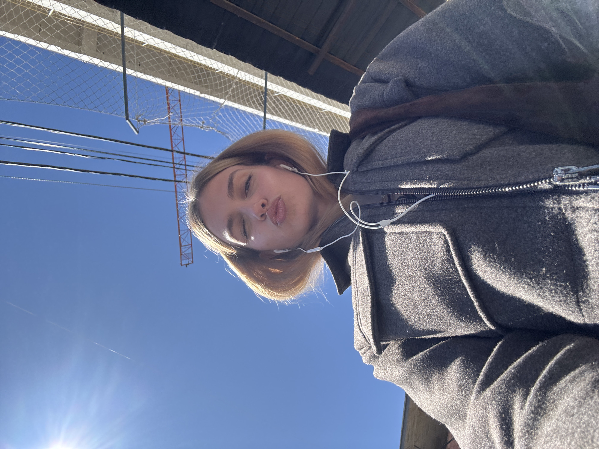 A woman wearing a gray coat and earbuds takes a selfie while standing outdoors on a sunny day. Above her, a chain-link fence and overhead structures, including what appears to be a crane, are visible against a clear blue sky.