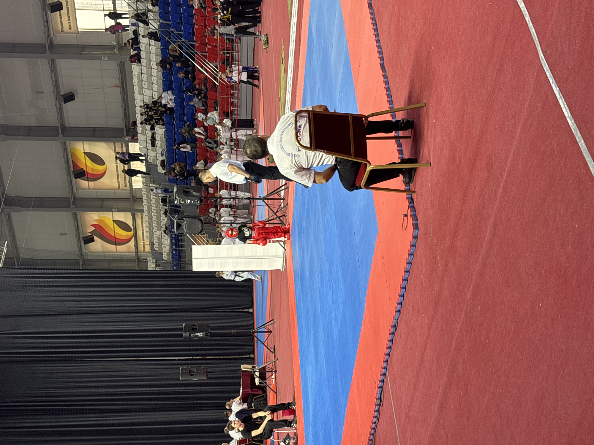 The image captures an indoor sporting event, possibly martial arts or a similar competition. The event appears to be held in a large arena, indicated by the tiered seating in the background filled with spectators, some wearing sports attire. The seating is patterned with red, white, and blue, the colors of the Russian flag. There is a large logo on the wall. Below the wall is a black curtain.
In the foreground, a red and blue mat defines the competition area. A referee or judge sits on a chair at the edge of the mat. A second person is wearing a white shirt.
Based on the writing on the logo, this appears to be in Perm, Russia.