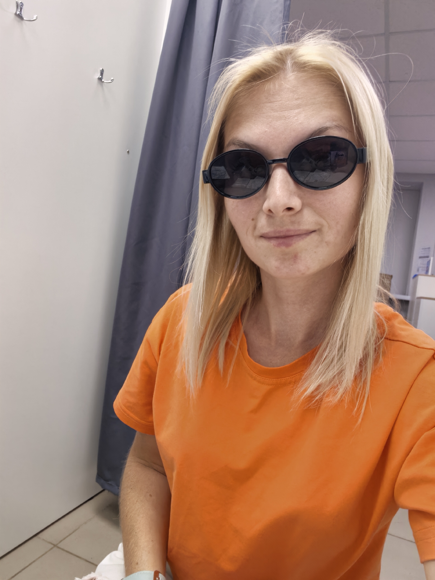 The image shows a blonde woman in an orange t-shirt and sunglasses, taking a selfie in what appears to be a fitting room or a changing area. She has blonde hair and is looking at the camera with a slight smile. The background shows a grey curtain on the right and a white wall with two hooks on the left. The floor is tiled. There are no visible elements in the image that would suggest it was taken in the city of Sterlitamak, Russia.