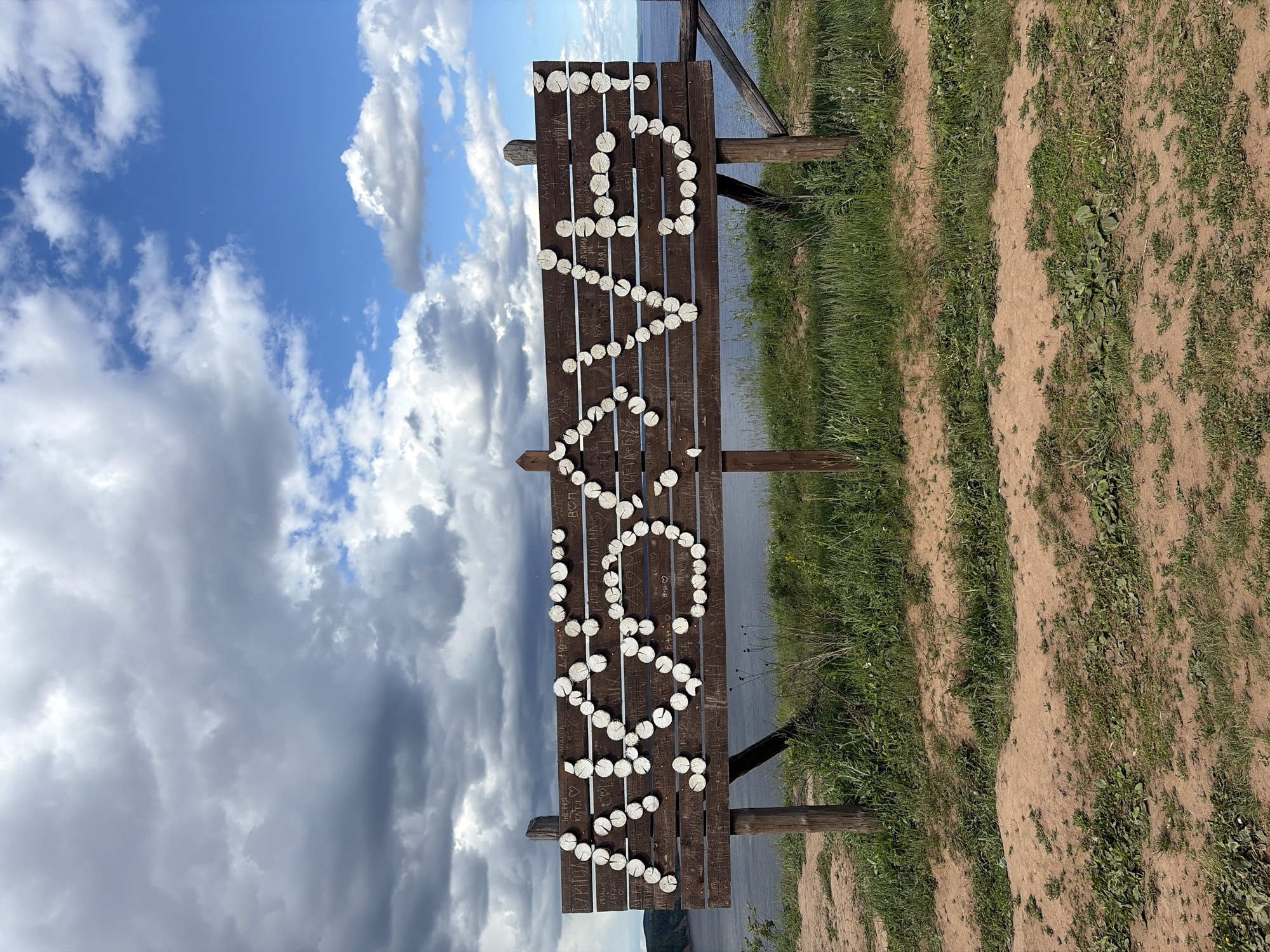 A landscape photograph shows a bench with the name of the city "Костоваты" formed by wooden slices. The bench overlooks a body of water under a sky with scattered clouds. The ground adjacent to the bench is a mix of dry dirt and green vegetation.
