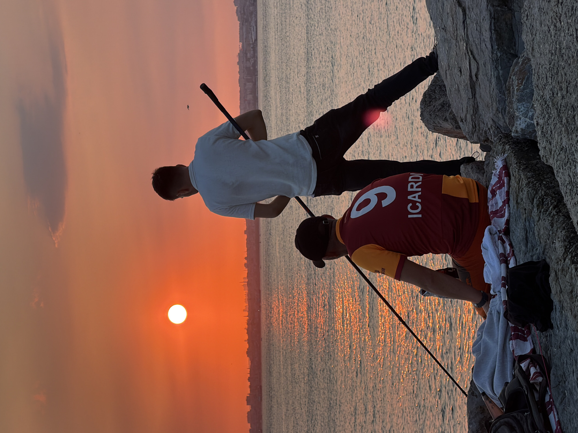 Two individuals are fishing on the shore of Kadıköy, Istanbul, Turkey, during sunset. The sun, a bright orange orb, is low in the sky, casting a warm glow over the scene and reflecting on the rippling water. The silhouette of the city skyline is visible on the horizon.
In the foreground, one person, wearing a red and gold jersey with the name "ICARDI" and the number "9" on the back, sits on a rocky surface with a fishing rod. They are wearing a dark baseball cap. Beside them are several items of clothing, including a gray blanket and a red and white striped towel. A backpack is also visible.
The second person stands behind the first, also holding a fishing rod. They are wearing a white polo shirt and dark pants. Their back is to the camera, and they appear to be looking out towards the water.
The rocky shoreline slopes down to the sea, and the water appears calm with gentle waves. The overall atmosphere is serene and peaceful, capturing a moment of quiet recreation against a beautiful natural backdrop.