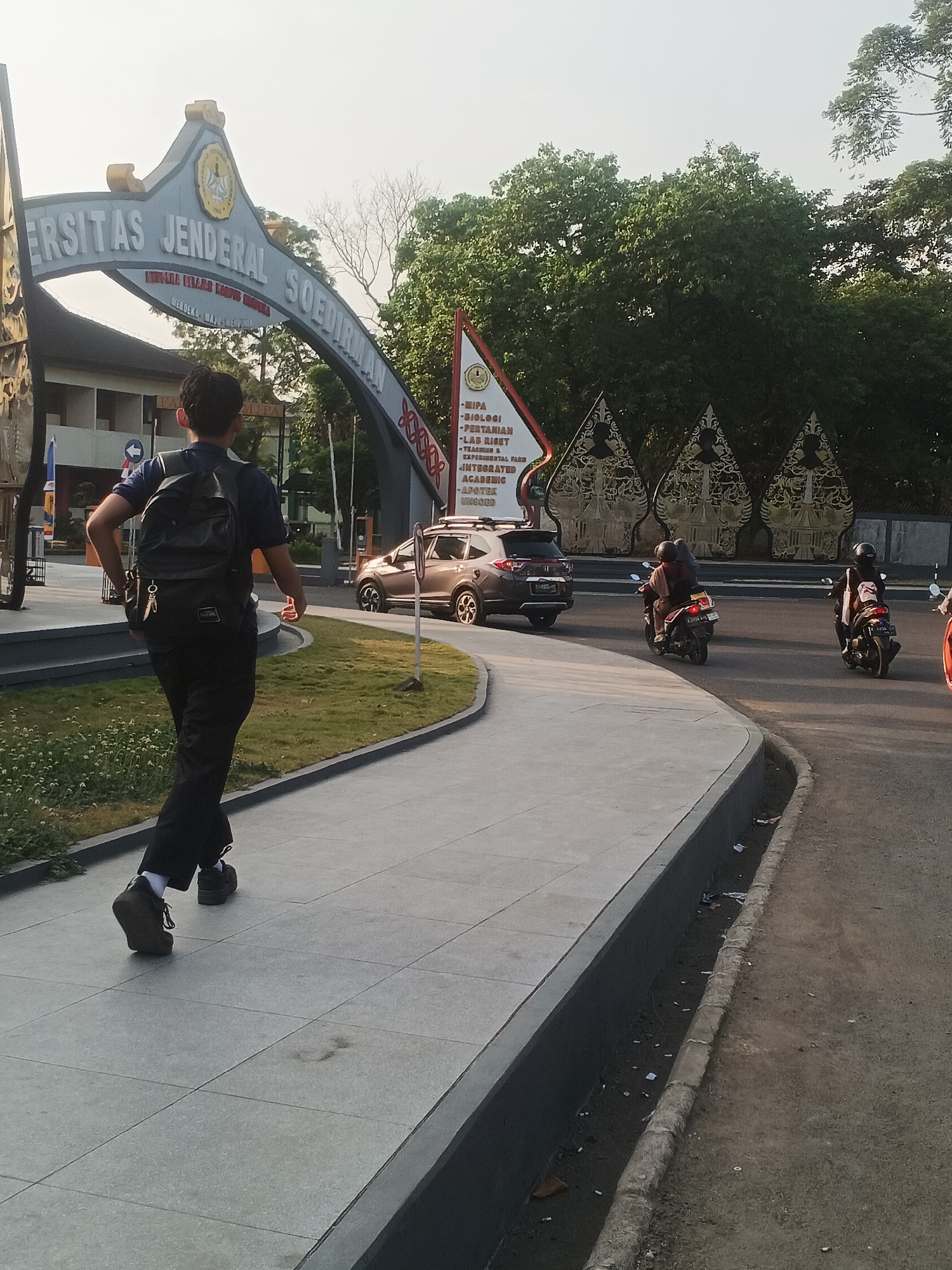The image depicts the entrance gate to Universitas Jenderal Soedirman in Karangwangkal, Indonesia. The gate is an archway with the university's name clearly visible. In the foreground, a young man wearing a dark blue shirt, black pants, and a backpack walks away from the camera on a paved walkway. He is on the left side of the walkway, moving towards the university entrance. To his right, a paved pathway curves to the right, bordered by a grassy area. Beyond the walkway, a road runs parallel to the entrance. Several vehicles are present on the road, including a silver SUV and two motorcycles carrying riders. The background features lush green trees and several decorative triangular structures. The lighting suggests it is daytime, possibly late afternoon given the long shadows.