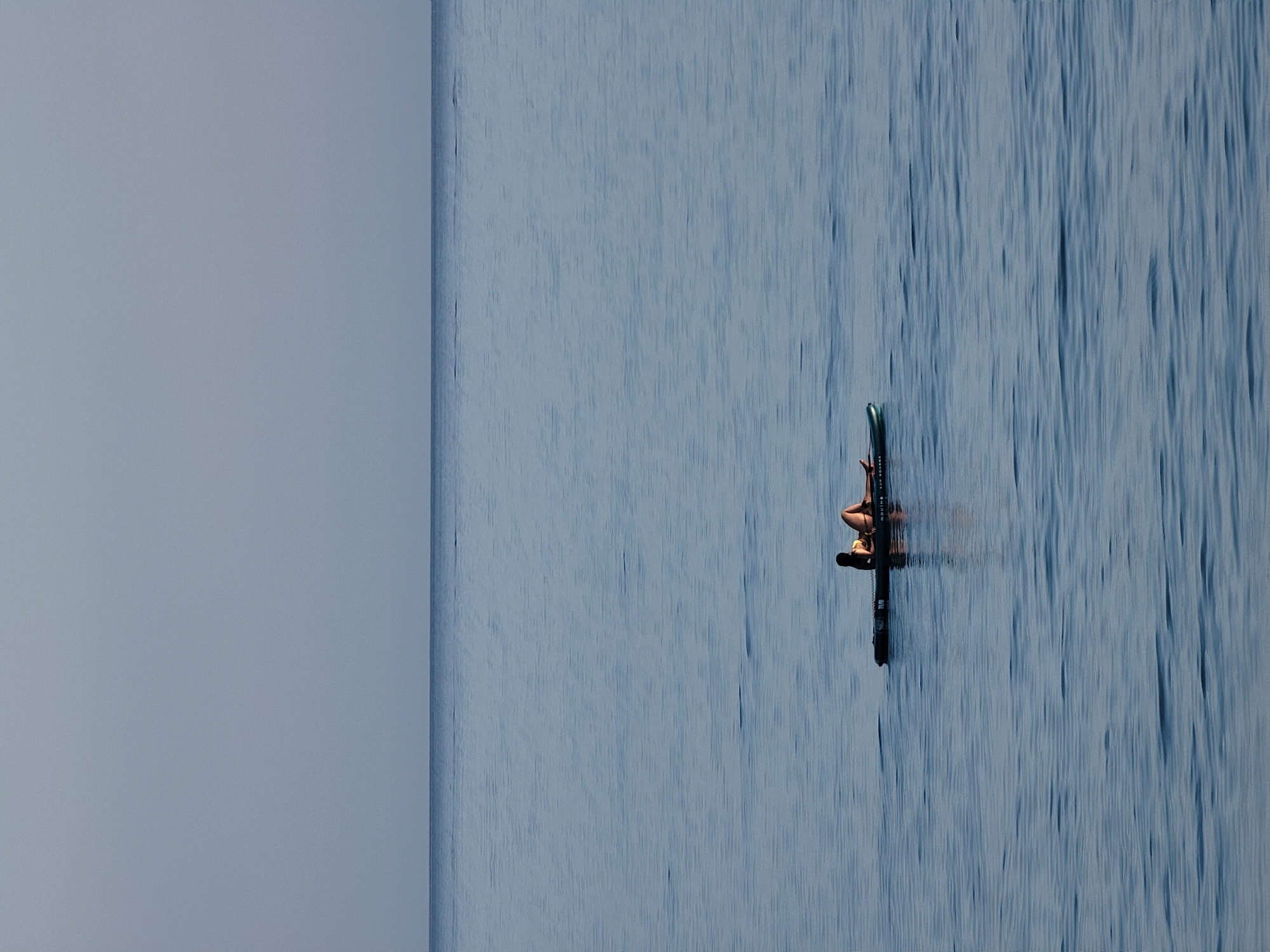 A person wearing a bikini is lying on a paddleboard in the ocean. The person is in the middle of the frame, to the left of center. The paddleboard is dark in color and appears to be floating on calm, blue water. The sky above is a pale, cloudless gray. The horizon line between the water and the sky is sharp and clear. The image was taken in the city of Quarteira, Portugal.