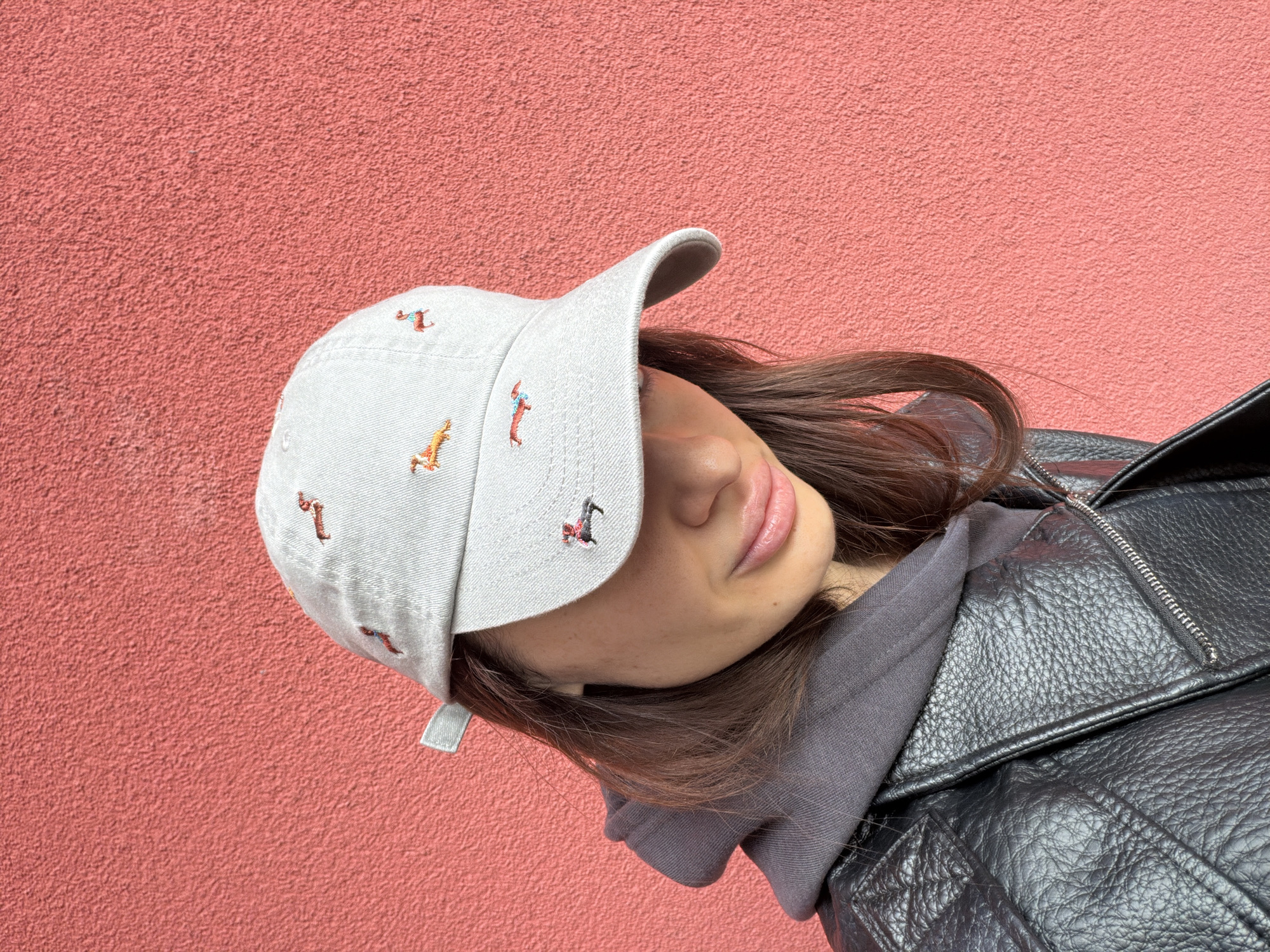 The photograph depicts a person wearing a light-colored baseball cap with embroidered figures, a grey hooded sweatshirt, and a black leather jacket. The person is positioned against a textured, salmon-colored wall. The image appears to be taken outdoors. Without additional information, I cannot confirm if the image was taken in Varna, Bulgaria.