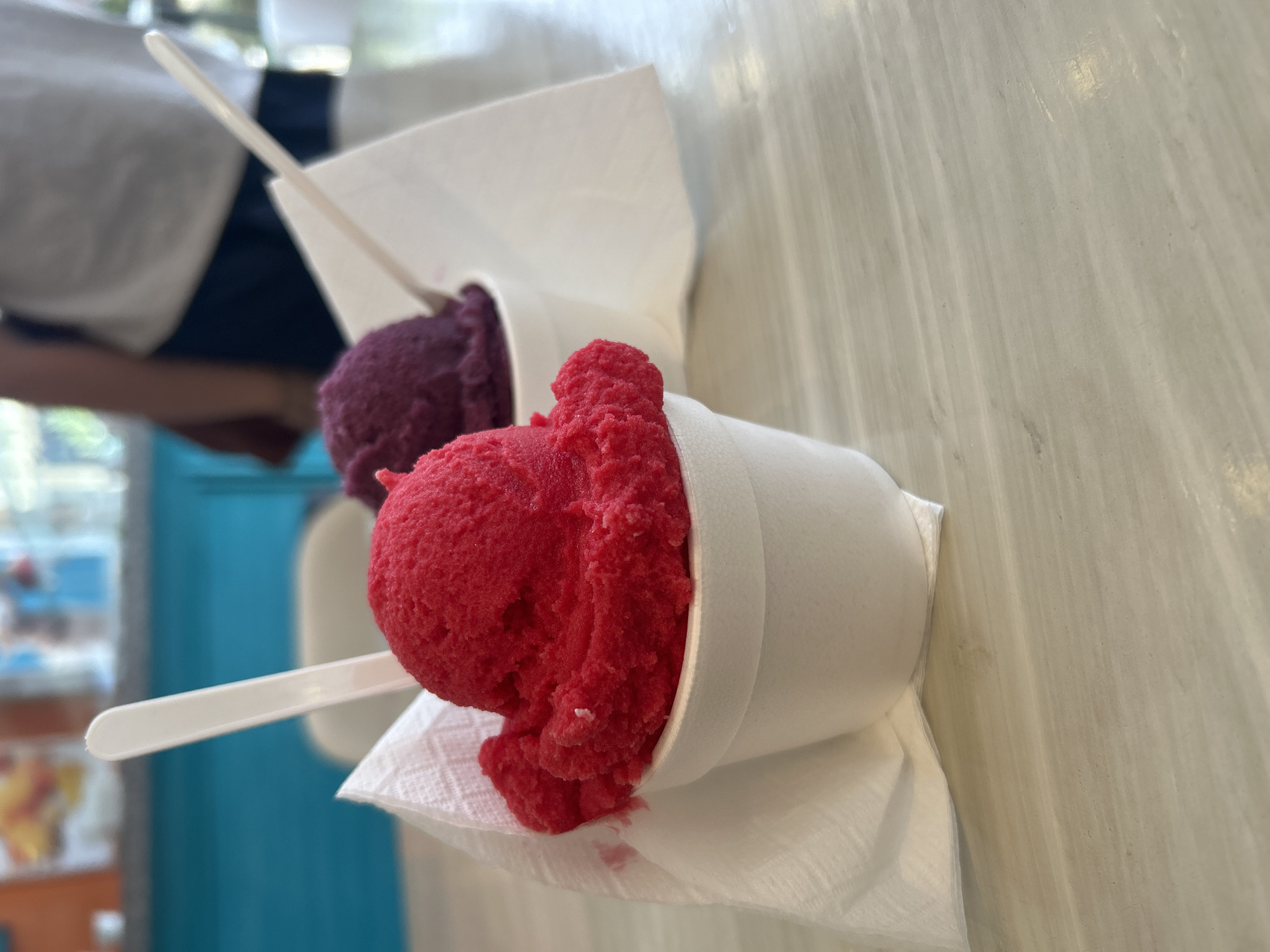 Two cups of shaved ice are on a white counter or table. The shaved ice is red and purple. The cups are white and have white napkins underneath them. There is a white plastic spoon in each cup. A person's arm is in the background wearing a white shirt and blue pants. There is a blue and orange wall in the background. The location is in the city of Unknown, Unknown.