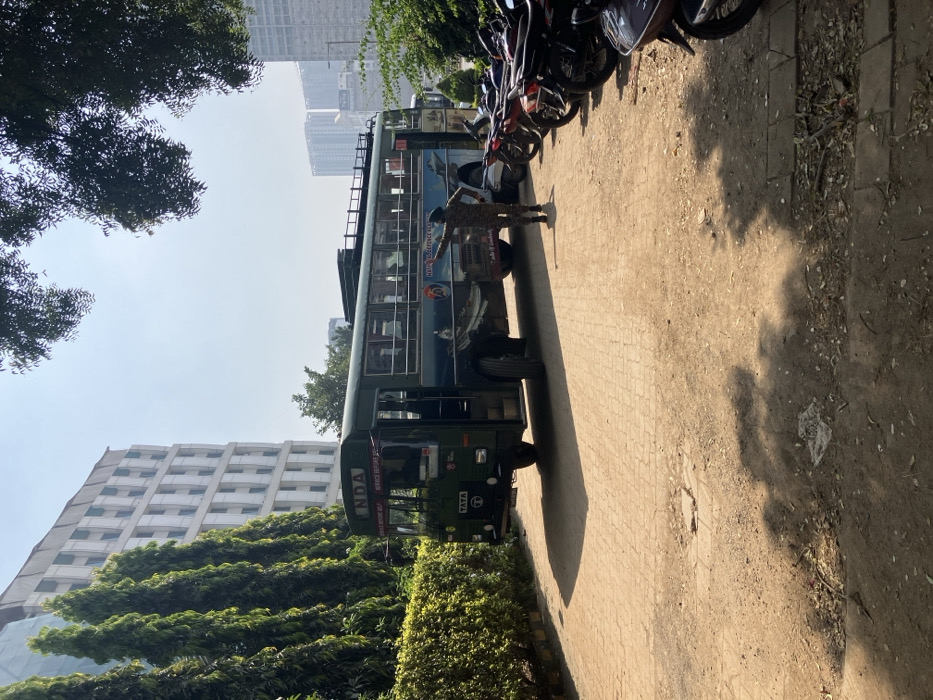 A medium shot captures a green NDA (National Defence Academy) bus, partially obscured by lush green bushes. A uniformed officer stands near the bus. Several motorbikes are parked behind it. The scene is set in an urban area with buildings visible in the background. The ground appears to be paved. The sunlight casts shadows, and the sky is a clear blue. The composition is slightly rotated.