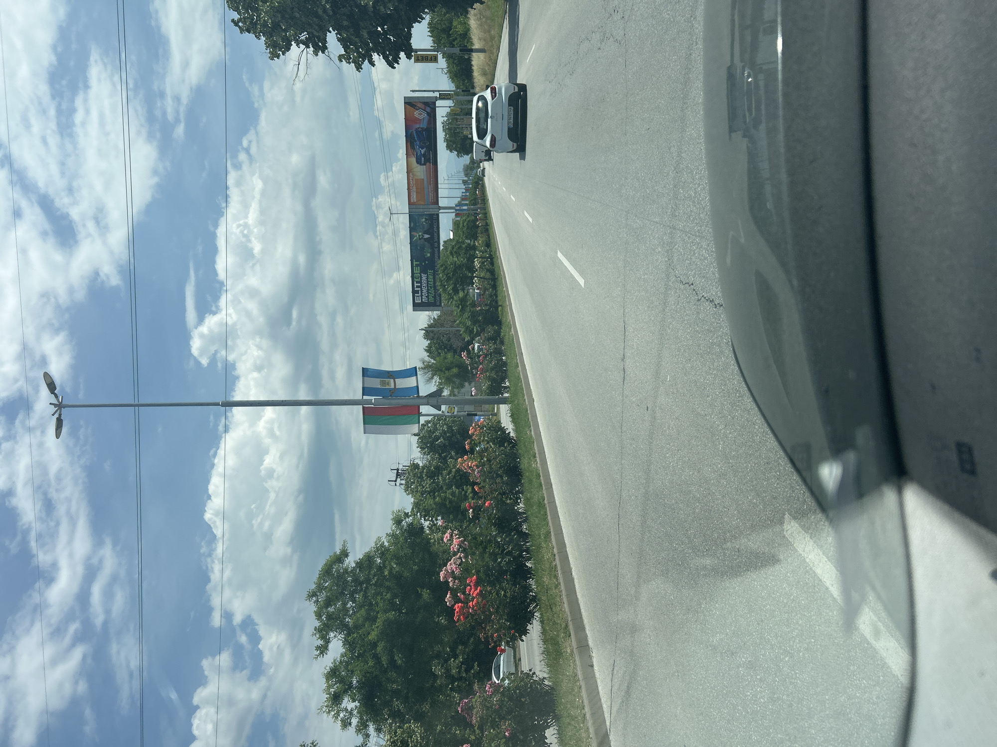 The image presents a daytime scene from the interior of a vehicle traveling on a multi-lane road in Plovdiv, Bulgaria. The road extends into the distance under a bright blue sky with scattered white clouds. A white car is visible ahead in the same lane.
To the left of the road, a tall street light pole displays two flags: the upper flag appears to be the blue and white flag of Plovdiv, featuring the city's coat of arms, while the lower flag is clearly the horizontal white, green, and red tricolor of Bulgaria. Lush green trees and bushes adorned with pink and red flowers line the roadside.
Several large billboards are prominent features along the route. One billboard prominently displays the text "ELITIBET" along with the Cyrillic phrase "ПРОМЕНЯМЕ ПРЕДСТАВИТЕ" (meaning "We change perceptions"). Another billboard advertises "RENAULT CAPTUR." Further down the road, a billboard for "EFFBET" is partially visible. Overhead power lines crisscross the sky. The image captures a sunny day, focusing on the road, commercial signage, and the distinct national and regional flags confirming the location.