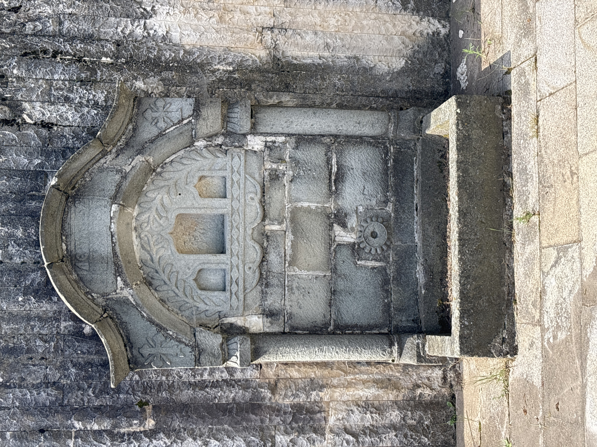 The image shows a stone fountain built into a rough stone wall. The fountain has an arched top with decorative carvings that include what appear to be two small windows or niches. Below the arch is a section of brickwork. In the center of the brickwork is a circular carving resembling a flower or sunburst. Below this is a stone basin that appears to be the reservoir of the fountain. The fountain is located on a paved area with some sparse vegetation growing around it. The wall behind the fountain is made of irregular stone blocks, some of which are covered in moss or lichen. The ground in front of the fountain is paved with large rectangular stone slabs. The overall impression is of an old, possibly historical, water feature. The fountain is located in Sofia, Bulgaria.