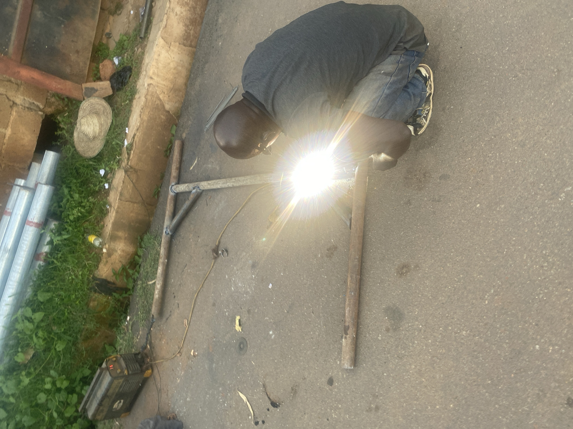 In Gura Top, Nigeria, a man is welding metal pipes on a paved street. He is wearing a dark t-shirt, blue jeans, and athletic shoes. The welding equipment, including a generator and welding rods, is positioned nearby. The scene is outdoors, with lush green vegetation and a concrete embankment visible in the background. Construction materials, such as more metal pipes and a straw hat, are scattered around the area. The bright light from the welding process illuminates the immediate vicinity.