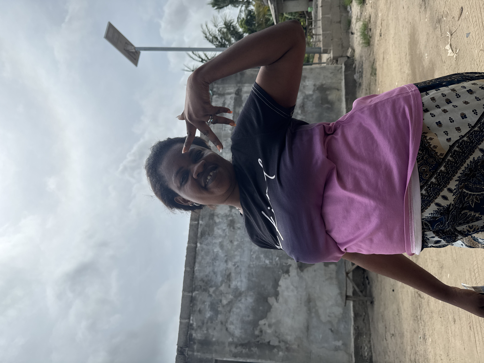 A woman stands outdoors on a dirt surface in Badagry, Nigeria. She wears a black and purple shirt, a pink top, and a patterned skirt. She smiles at the camera with her left arm bent upwards, fingers spread. A concrete structure with a small opening is behind her. A solar-powered streetlight post stands to the left. The sky is overcast. Vegetation is visible in the background.