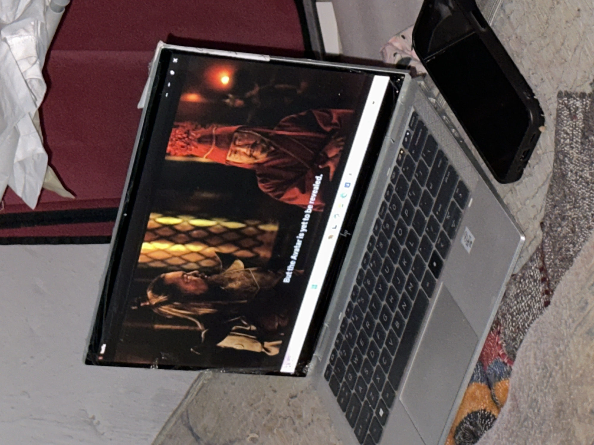 A laptop rests on a surface that appears to be part of a rug or possibly a textured fabric. The laptop screen displays a scene with two characters, one clad in red and facing upwards, the other with long hair lying on their back. Text on the screen reads "But the Avatar is yet to be revealed". A black object is positioned to the right of the laptop. The laptop's keyboard is visible, and the keys are in a typical QWERTY layout. The background contains hints of a red surface and some white cloth, but the overall scene lacks clear environmental context. Kirana, Nigeria is not visible in the photo.