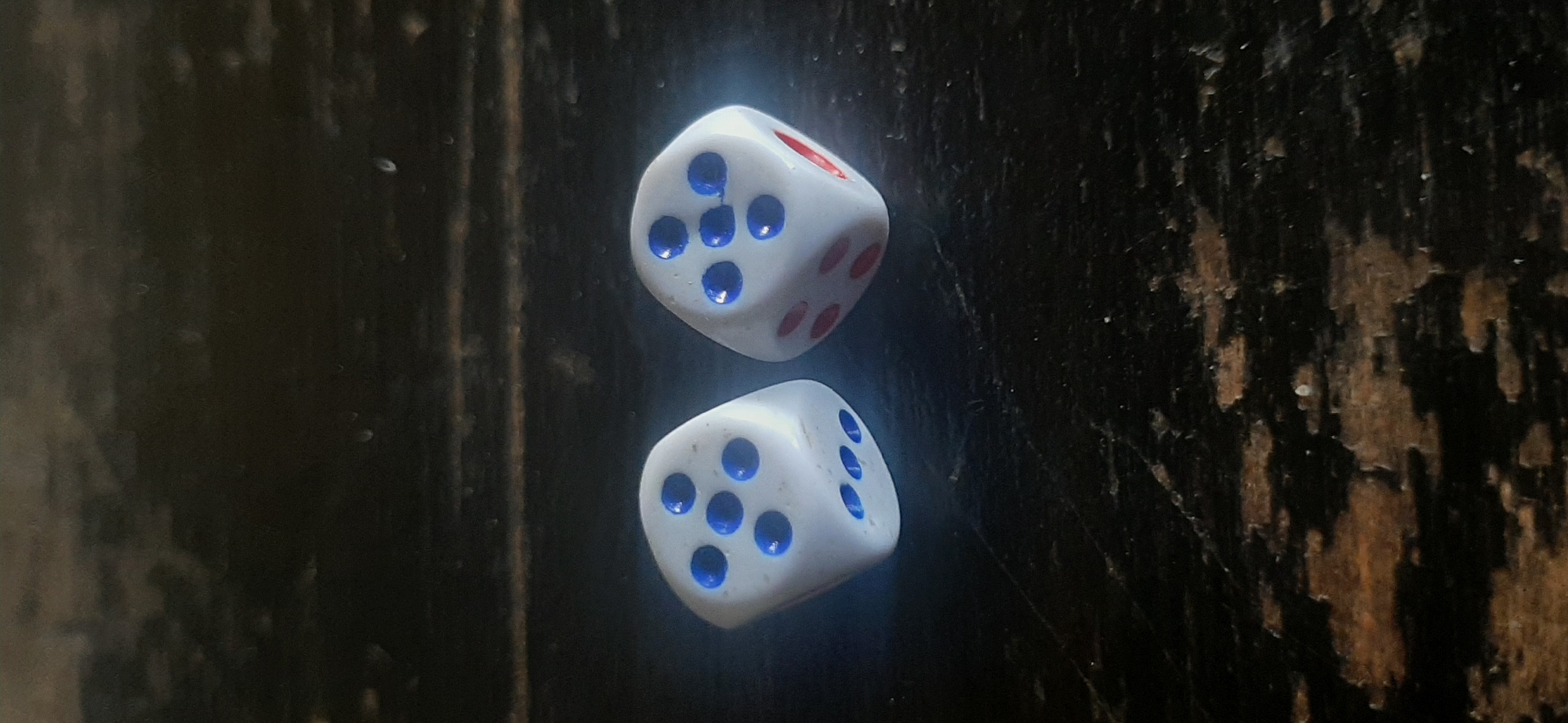 Two white dice with blue pips are positioned vertically against a dark, textured background. The top die shows a face with five pips and parts of adjacent faces are visible, revealing a face with two pips and a face with one red pip. The bottom die displays a face with five pips and portions of adjacent faces are also visible, showing faces with three pips and one pip. The lighting highlights the pips on the dice and casts subtle shadows. The background appears to be a rough, dark surface, possibly wood with an aged or distressed finish, with vertical streaks and variations in tone. The scene is a close-up, focused on the dice. No people, text, or specific time of day or weather cues are discernible. The overall impression is still and focused on the objects.