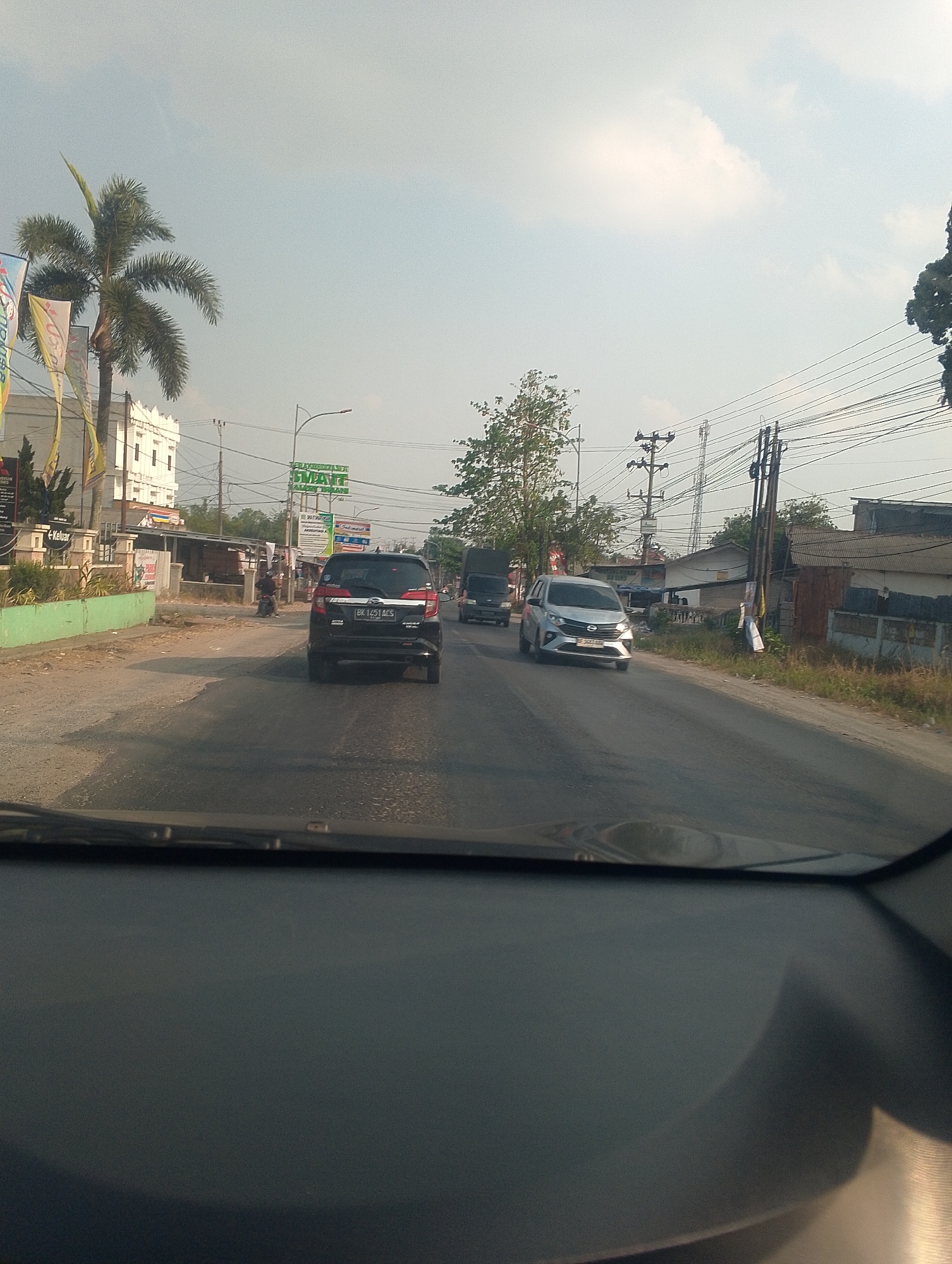 The image captures a street scene in Bandar Jaya, Indonesia, from the perspective of a vehicle's interior. The road ahead is paved, with traffic including a black SUV, a silver sedan, and a dark truck. Buildings and businesses line the right side of the road, featuring signage that includes "SMART" in green text. A tall palm tree is visible on the left side, near a white building. Power lines and utility poles are prominent features of the environment. The sky is clear with some light clouds, suggesting daytime conditions.