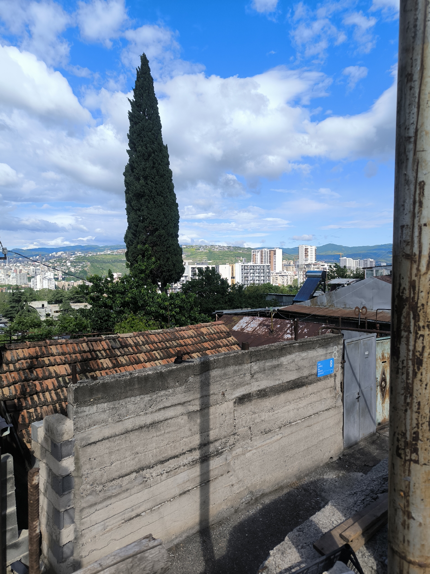 The image shows a view from a slightly elevated position in Tbilisi, Georgia, overlooking a section of the city. In the foreground, there is a concrete wall and a dilapidated fence with a metal door, indicated with number 8 blue sign. Behind the wall, there are several buildings with red-tiled roofs and a tall, slender, dark green cypress tree. In the background, there is a cityscape with several high-rise buildings and a mountain range in the distance. The sky is partly cloudy with patches of blue. The photo is taken during the daytime. A rusty pole is present on the right side of the image.