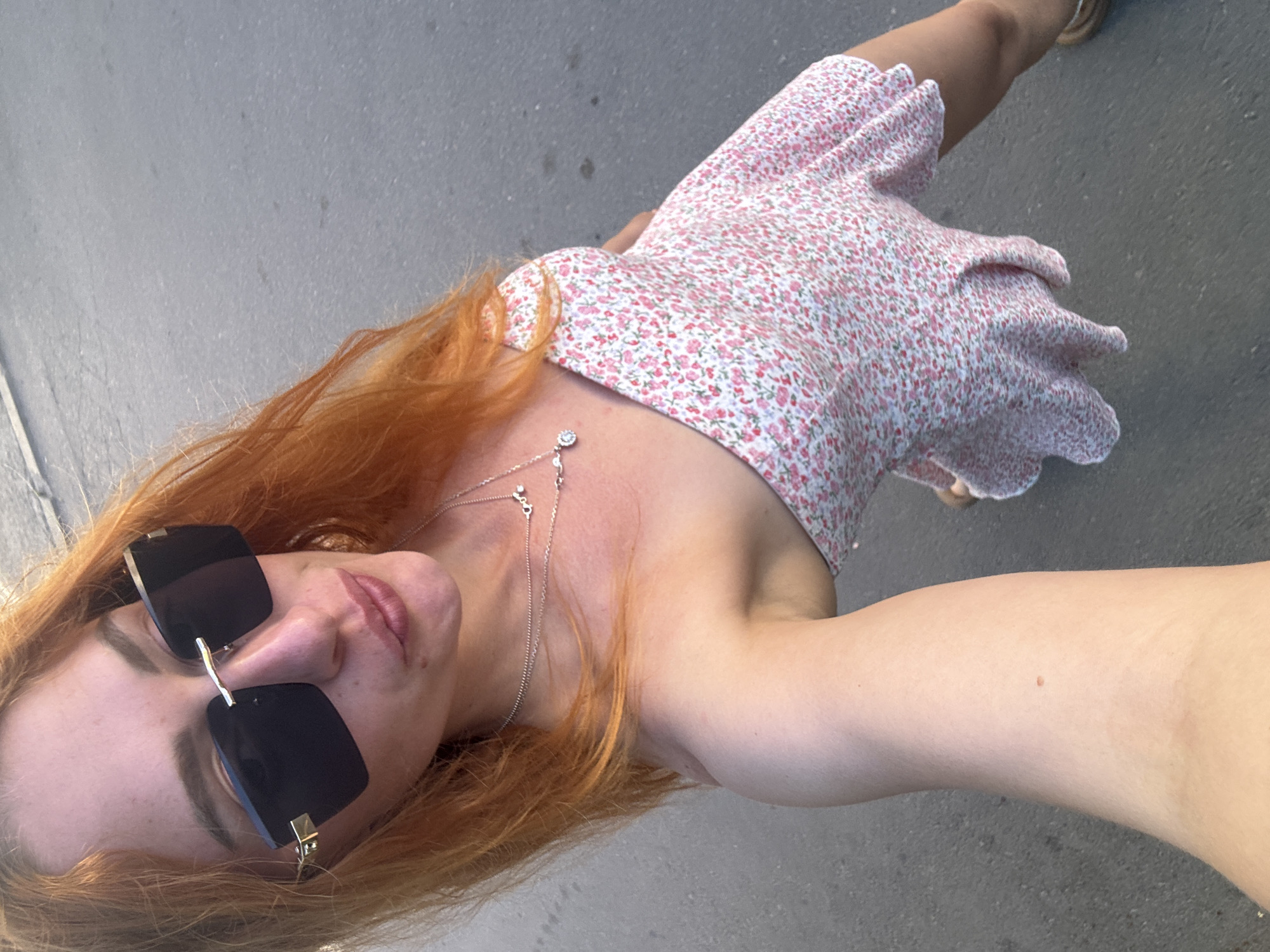 A high-angle selfie captured outdoors depicts a woman with long, wavy, reddish-orange hair. She wears large, dark, rectangular sunglasses featuring gold-colored accents on the bridge and temples, obscuring her eyes. Her lips are slightly pursed. She is adorned with two layered silver-colored necklaces, one featuring a small round, faceted pendant and the other an abstract, possibly script-like, pendant. The woman is wearing a strapless or off-the-shoulder short dress patterned with small pink, red, and white flowers on a white background. Her left arm, holding the camera, is visible in the lower-left corner, and her right leg with a light-colored sandal is partially visible on the right. The background consists of uniform gray asphalt or pavement, indicating an outdoor setting under bright natural light. The image is described as being in the city of Budva, Montenegro, although no specific landmarks are discernible within the frame to confirm this location.