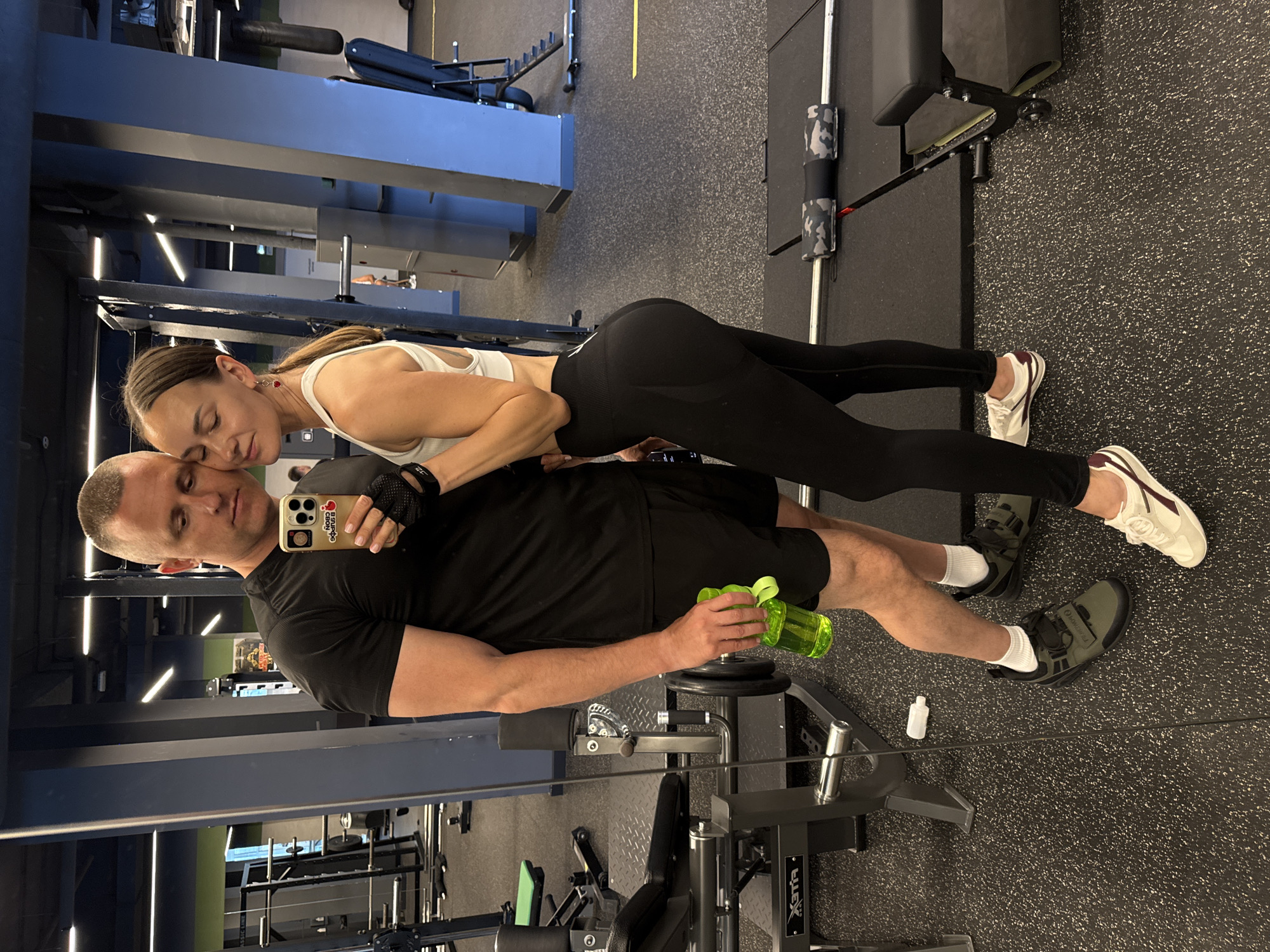 A man and a woman, appearing to be a couple, pose for a selfie in a gym. The man, with a buzz cut, wears a black t-shirt, black shorts, and olive green athletic shoes. He holds a bright green water bottle in his right hand. The woman, with her hair in a ponytail, wears a white sports bra and black leggings. She has red earrings. They are standing close together, with the woman's head resting on the man's shoulder, and both have their eyes closed. The background shows gym equipment, including weight racks, benches, and weights, all set against a dark grey and blue color scheme. The floor is black rubberized gym flooring. The lighting is bright, likely from overhead gym lights, casting minimal shadows. The overall scene is captured in a mirror, indicating a reflection. The context of Sochi, Russia, is not explicitly depicted in the image itself, but is provided as a locational detail.