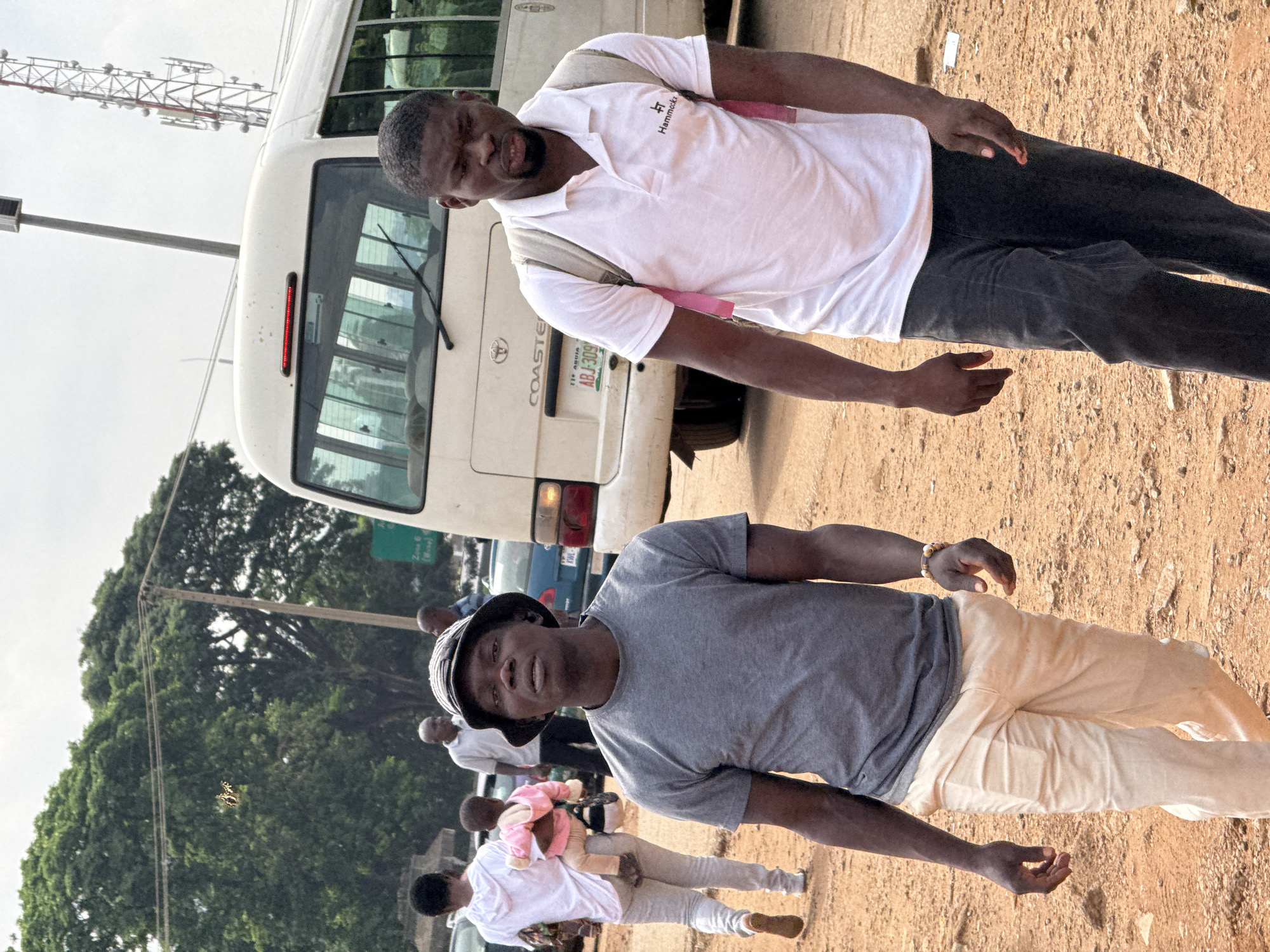 Two men are walking in a dusty, outdoor area in Abuja, Nigeria, with a white minibus and a cell tower visible in the background. The man on the left, wearing a grey t-shirt and light-colored trousers, is looking forward. He has a bucket hat on his head and a bracelet on his right wrist. The man on the right, wearing a white polo shirt and dark trousers, is also looking forward. He has a backpack with pink straps. In the background, a woman is carrying a child, and other people are standing near vehicles. The ground is covered in reddish-brown dirt.