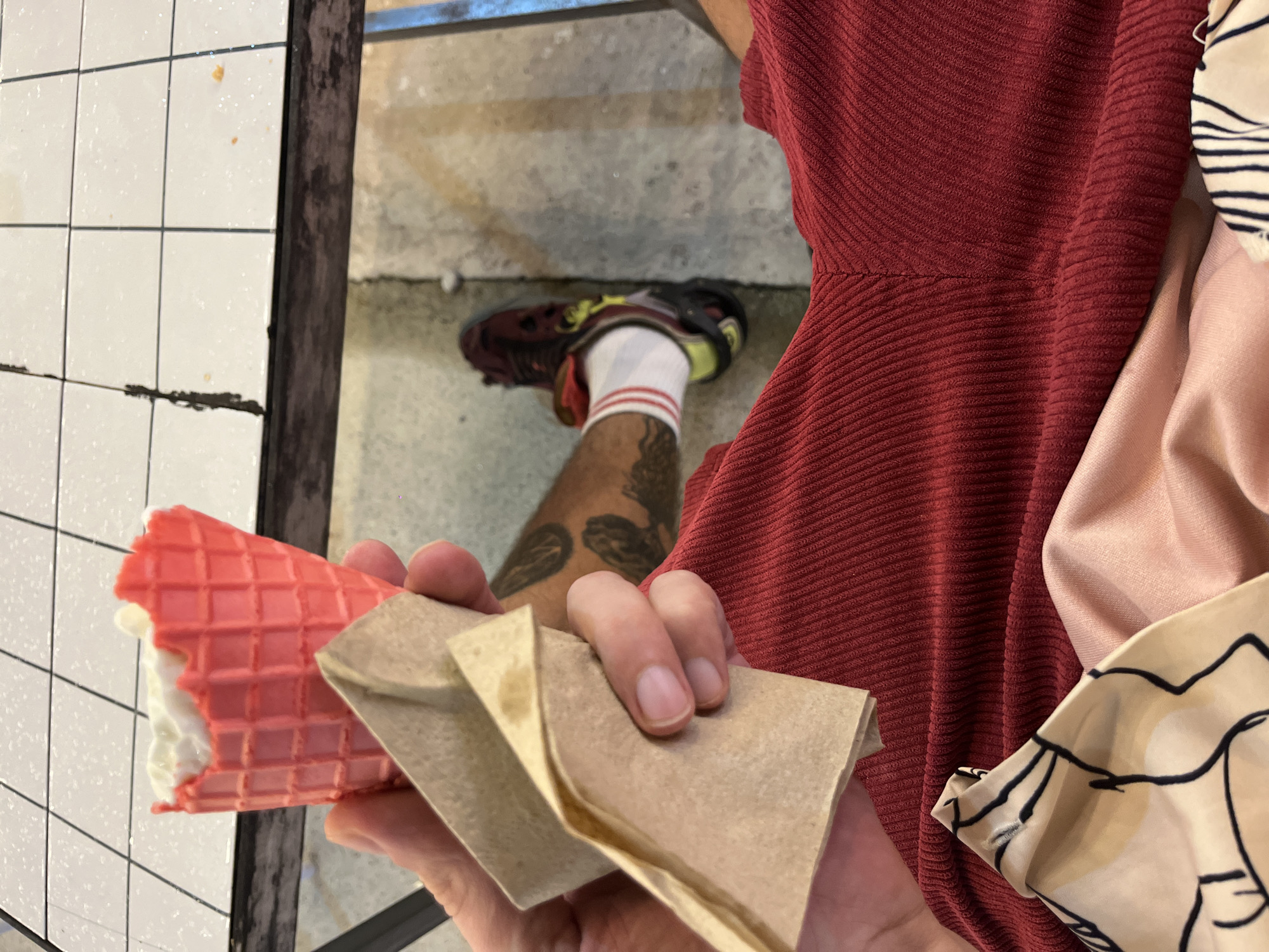 In the city of Phuket City Municipality, Thailand, a person is holding a partially eaten pink ice cream cone and a crumpled brown napkin. The person is wearing red shorts and white socks with red stripes, and a black and yellow shoe. The background shows a tiled surface and a concrete floor. The image is taken from a close-up perspective, focusing on the ice cream cone and the person's hand.