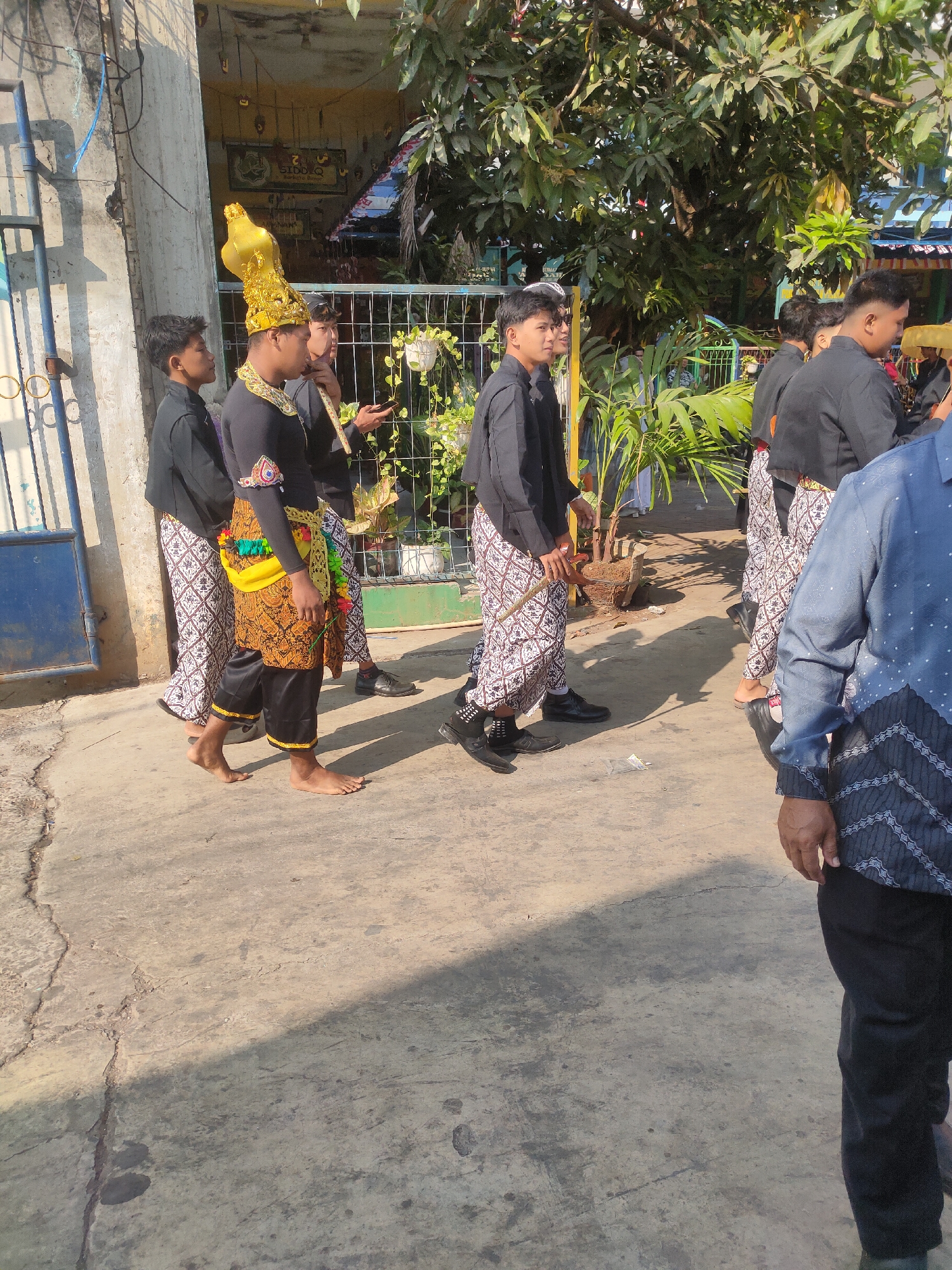 A group of young men in traditional attire are walking on a paved street in Payaman, Indonesia. The central figure wears an elaborate gold headdress and a black long-sleeved shirt with colorful trim on the sleeves and waist. He is also wearing patterned pants and a yellow sash. Other young men are dressed in black shirts and patterned pants. They appear to be participating in a procession or cultural event. The environment includes a building with a blue gate on the left, lush green foliage in the background, and a fence. The ground is made of concrete with some cracks. The lighting suggests it is daytime.