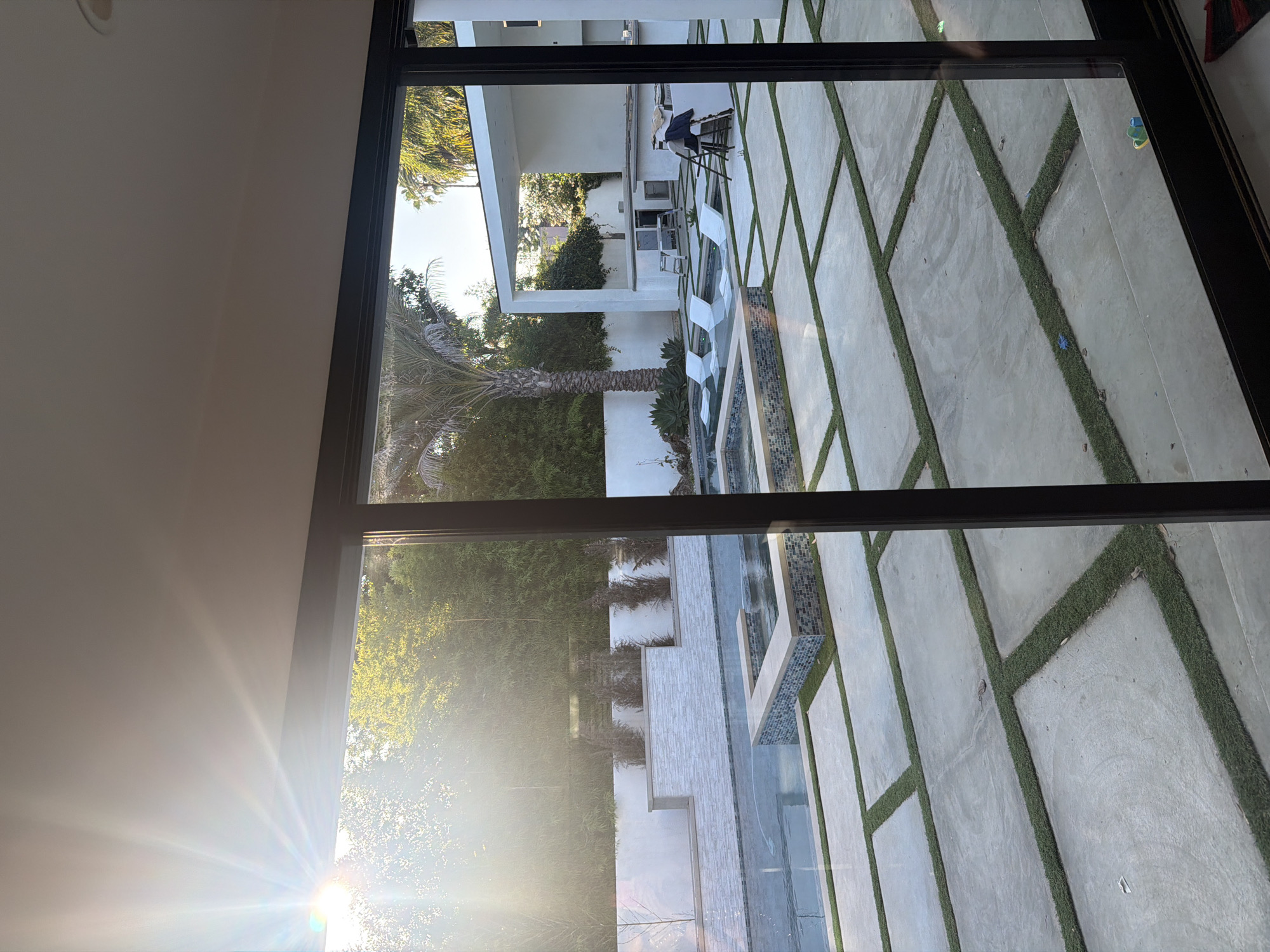 Here's a description of the image:
The photograph presents an outdoor scene, likely captured through a large window or glass door. The setting appears to be a modern backyard or courtyard. The foreground is occupied by the window frame. Beyond the frame, a geometric pattern of concrete pavers separated by strips of grass forms the main surface.
The courtyard features a shallow water feature with stone borders and white lounge chairs. There's a covered outdoor dining or lounging area with chairs and a small bar area set against a white wall with climbing greenery. In the background, a dense arrangement of trees and a palm tree is visible.
The location is a residential property located in Los Angeles, California, United States of America.