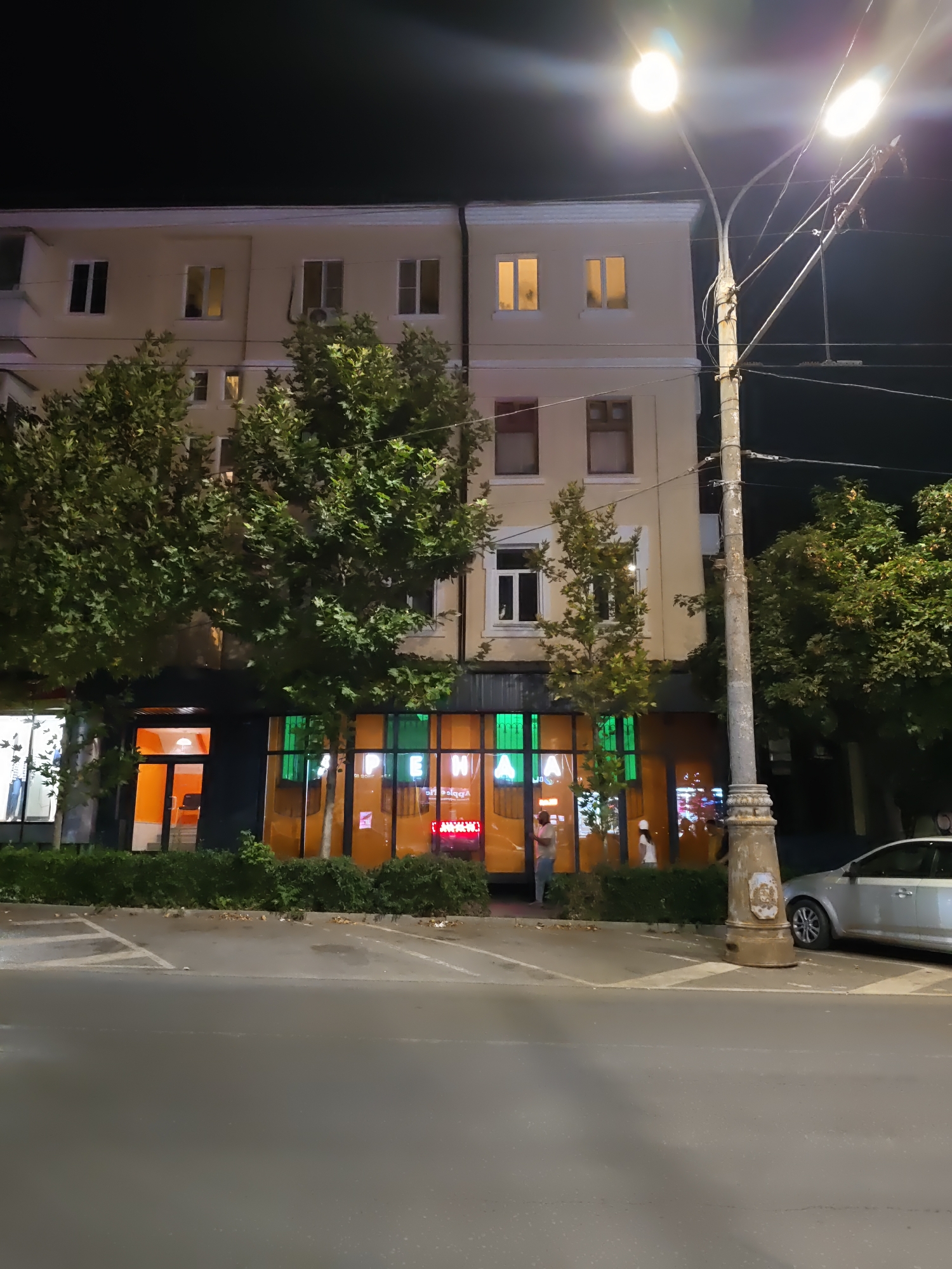 An exterior nighttime shot captures a light-colored building with several windows, some illuminated from within, alongside a street and sidewalk. A mature tree with dense green foliage partially obscures the lower facade of the building. The ground level features a storefront with bright orange glass and green-tinted windows, revealing interior lights and signage. Two individuals are visible near the storefront entrance. A lamppost with two bright lights stands prominently on the sidewalk, illuminating the scene. A silver car is parked on the right side of the image. The scene is located on a street in the city of Rostov-on-Don, Russia.