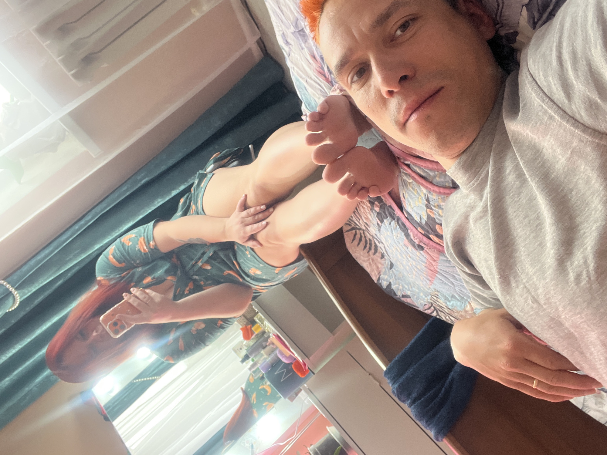 A selfie-style shot depicts a man lying on a bed, with a woman lying on the bed in the background. The man has light skin, short dark hair with orange dye at the roots, and is wearing a gray long-sleeved shirt. His right hand is visible, showing a gold ring on his ring finger. Parts of his right foot are also in the shot. The woman in the background has long red hair and is wearing a patterned top and shorts. She is holding a phone. The bed has a patterned bedspread with shades of pink, blue, and gray. A dark teal curtain hangs in the background. A shelf with various items is visible. The location is Unknown, Unknown.