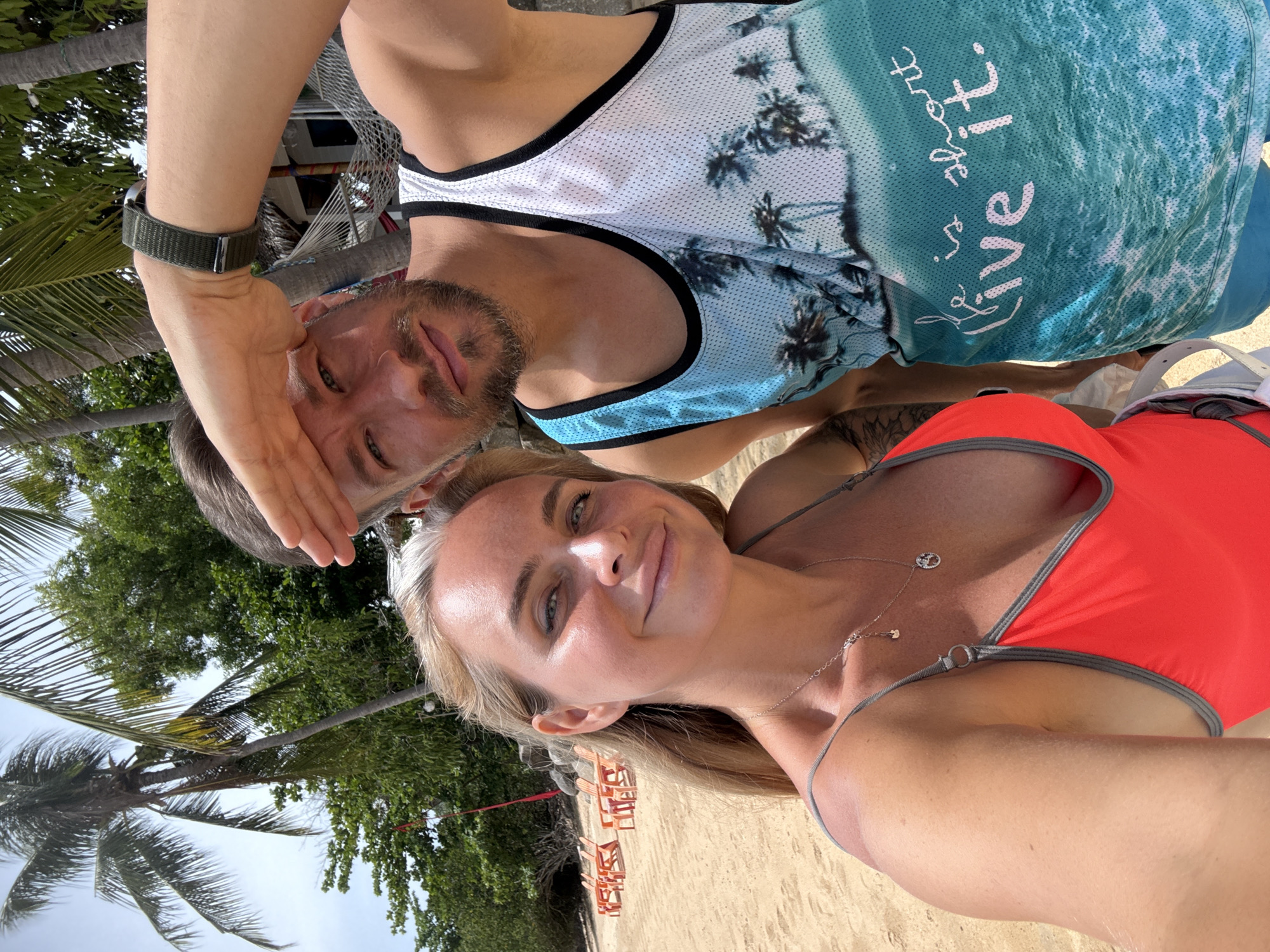 A close-up, selfie-style shot captures a couple on a sandy beach in Baan Bo Phut, Thailand. The woman, in the foreground on the left, has blonde hair and is wearing a red swimsuit with grey trim and a silver necklace. She smiles at the camera. To her right, a man with short brown hair and a beard shields his eyes with his hand. He is wearing a white tank top with a tropical palm tree print and the text "Life is short, live it." visible on the front. Palm trees and lush green foliage are visible in the background, with several orange beach chairs set up on the sand. A hammock is also visible in the distance. The lighting suggests a bright, sunny day.