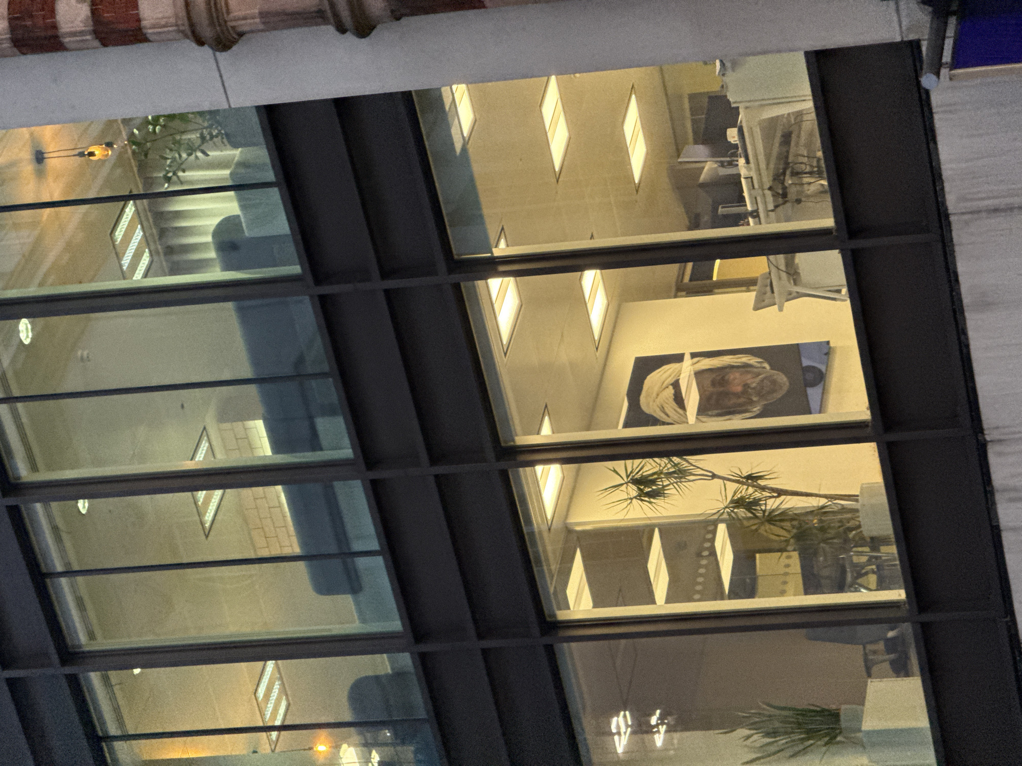 The image shows a view through a multi-paned window into an office space. Within the office, fluorescent lighting is visible, along with office furniture including desks and chairs. A large portrait is displayed on one of the office walls, alongside a potted plant with spiky leaves. The window frames are dark in color. Through the reflections in the glass, we can see the ceiling and lighting of the room where the photo was taken, including what appears to be a brick facade at the top of the image. Given the architecture and general ambiance, this photograph was likely taken in a commercial or business district of London, United Kingdom.