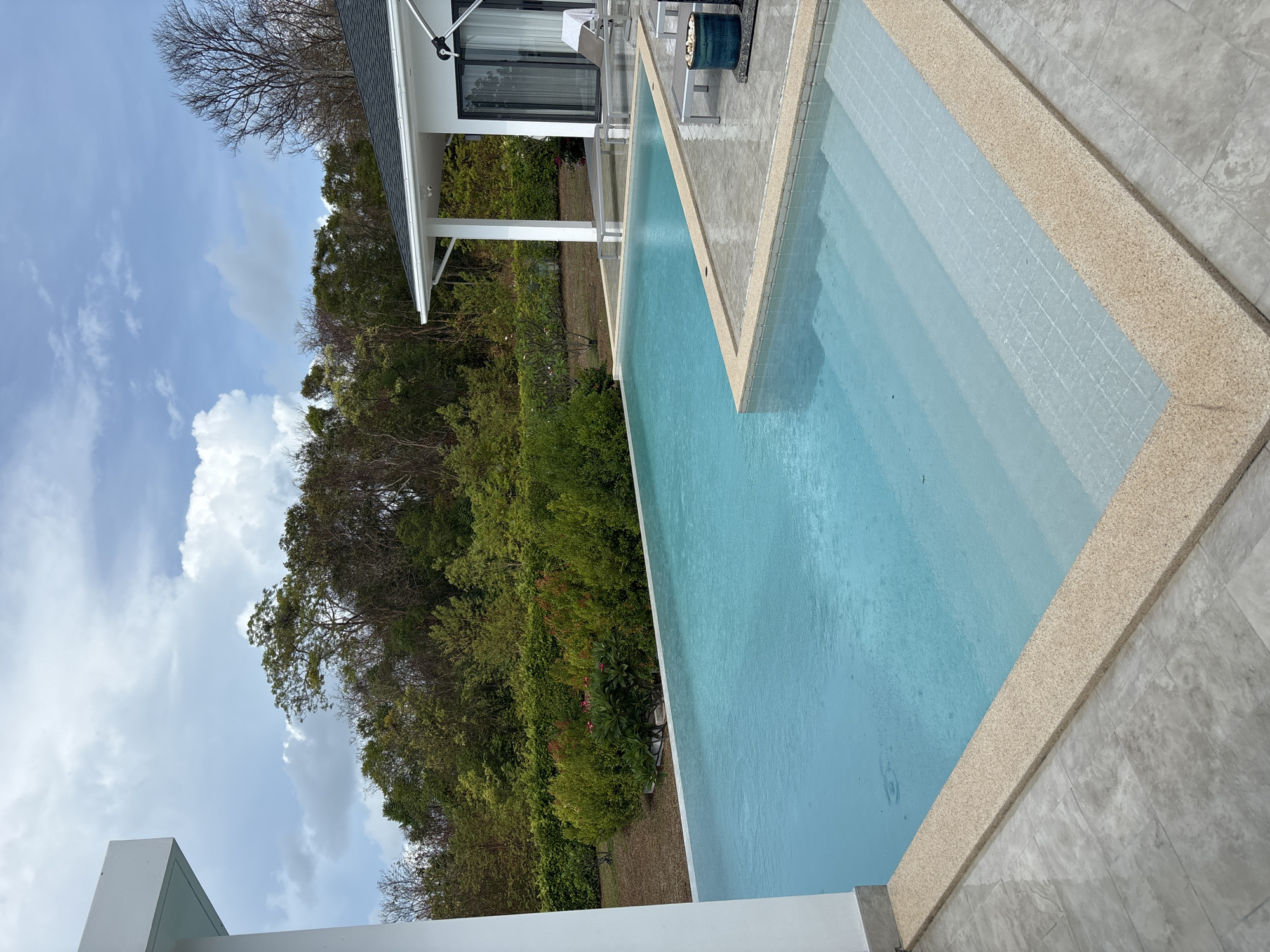 The image shows a swimming pool located in Ban Bang Khao, Thailand. The pool has a light blue color with steps leading into it. The surrounding area is decorated with light brown tiling. A building with a white exterior is situated next to the pool, partially obscured by lush green trees and vegetation. The sky is visible in the background, featuring scattered clouds and a hint of blue.
