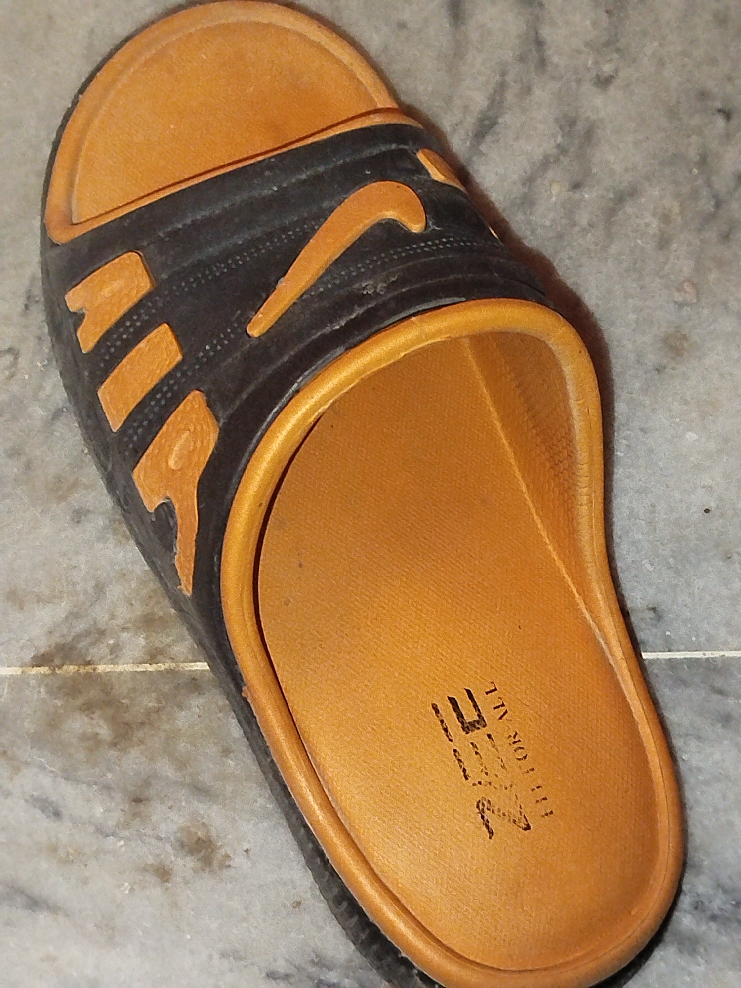 A single brown and black slide sandal is positioned on a gray and white marble floor. The sandal has a tan sole with a textured pattern. The strap of the sandal is black with three orange stripes on the side and a prominent orange Nike swoosh logo. The words "FIT FOR ALL" are embossed in black on the inner sole of the sandal, along with a logo that is difficult to decipher. The background is a marble floor, indicative of an indoor setting, possibly a home or a shop. The lighting suggests it is an indoor environment. There are no people or other objects present in the immediate vicinity of the sandal. The image is taken from a slightly elevated angle, looking down at the sandal. No specific location in Lassan, Pakistan can be determined from the visual information provided.